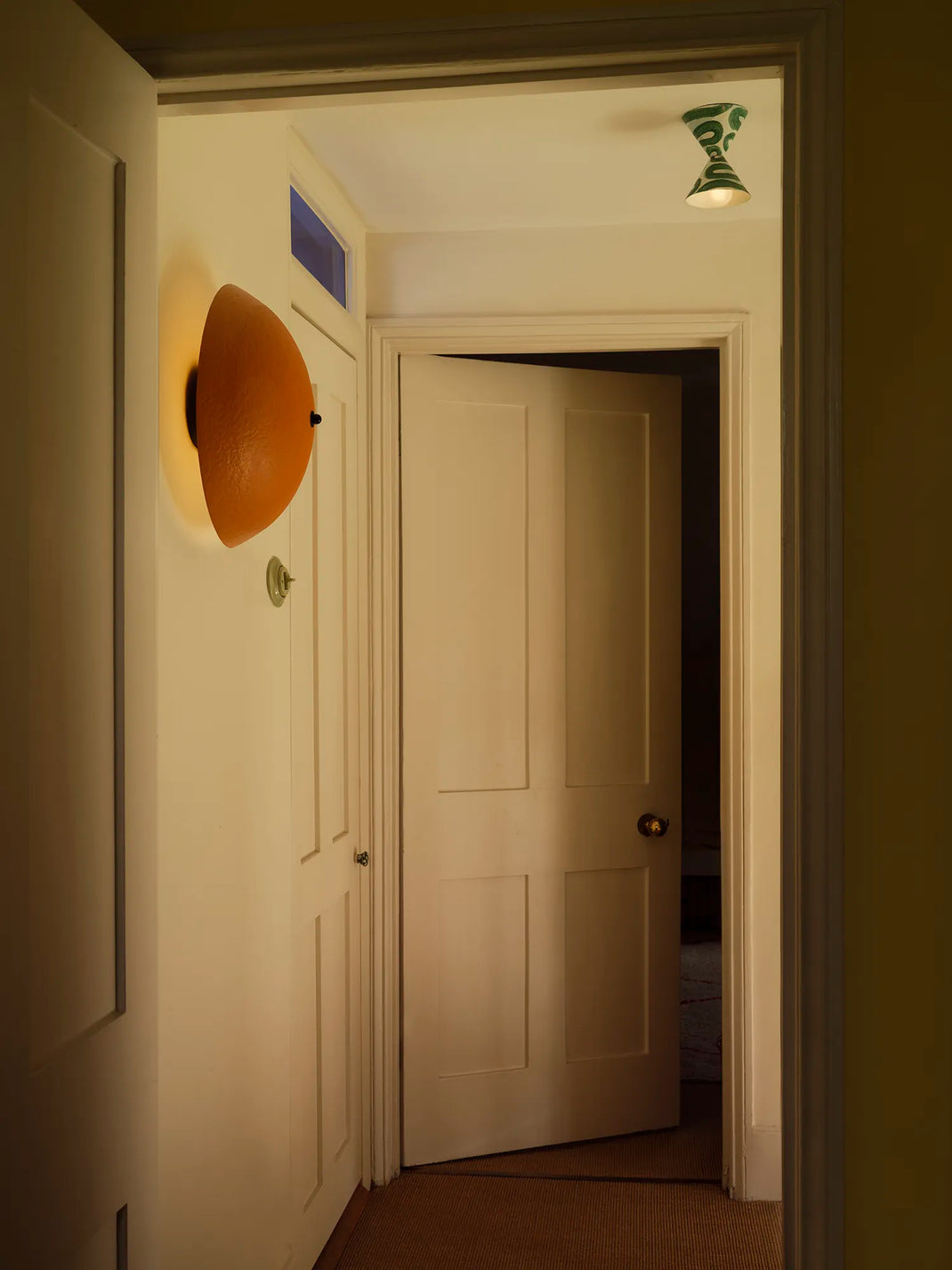 A warm, dimly lit interior hallway with cream-colored paneled doors, a textured orange wall sconce on the left, and a green decorative wall fixture on the ceiling.