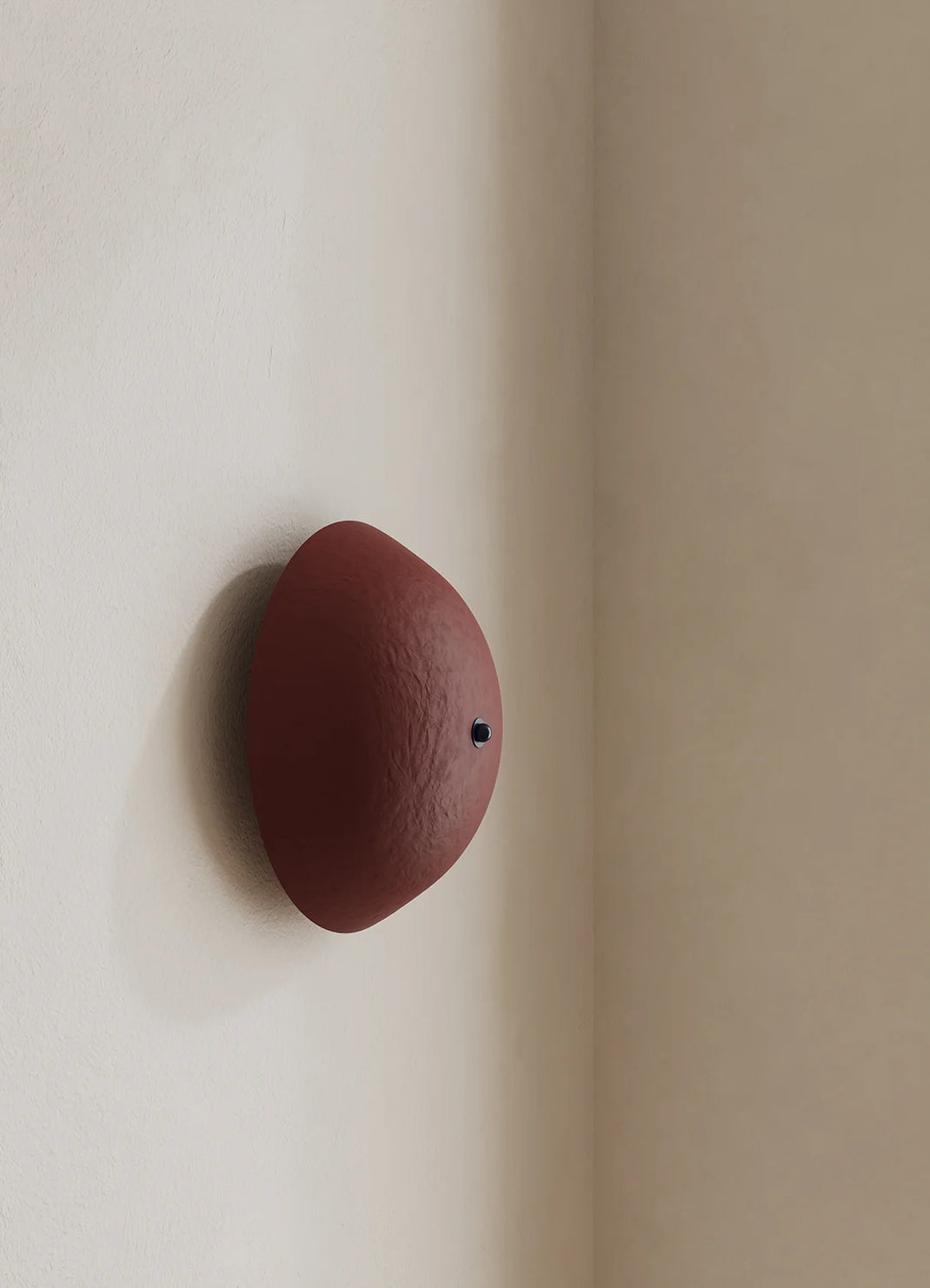 A minimalist wall sconce in a matte deep red finish, mounted on a warm beige plaster wall corner.