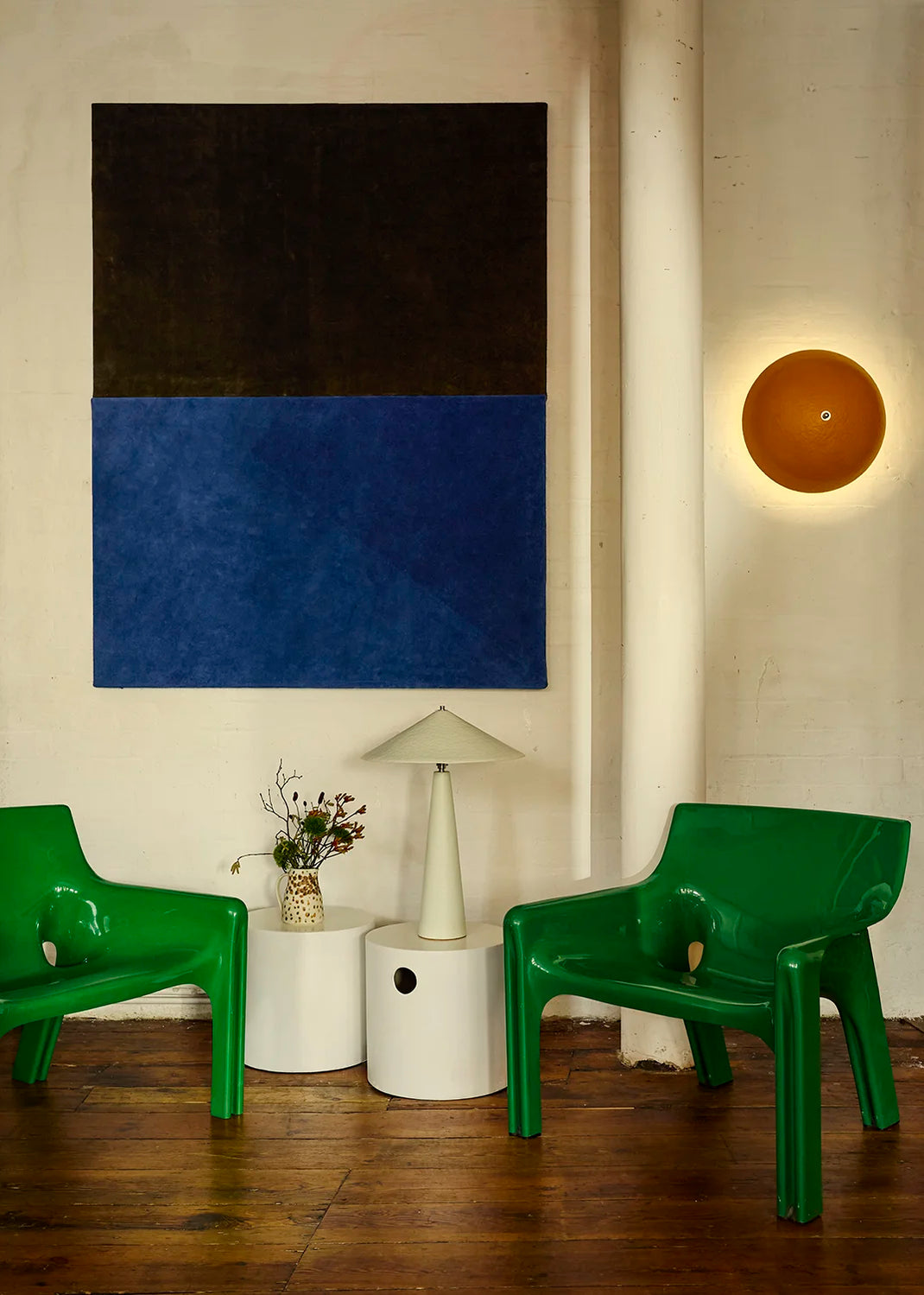 A styled interior scene with two bright green molded plastic chairs, a white side table, a white table lamp, and a bright orange wall sconce backlighting the wall.
