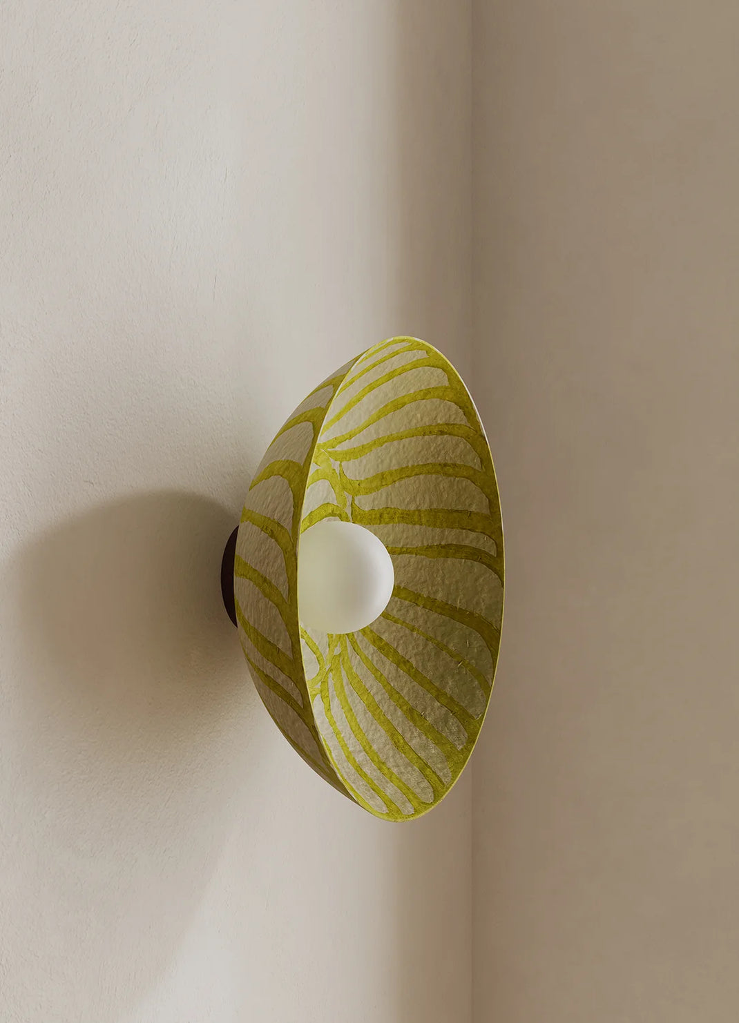 A modern wall-mounted sconce with a conical shade featuring a yellow and white wavy pattern, shown lit against a warm beige corner wall.