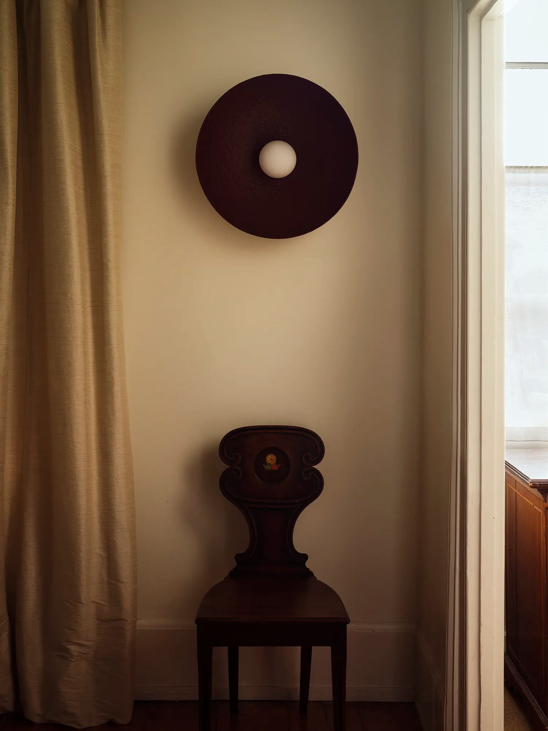 A dark red circular wall sconce with a white globe bulb, mounted on a beige wall above a dark wood chair in a room setting.