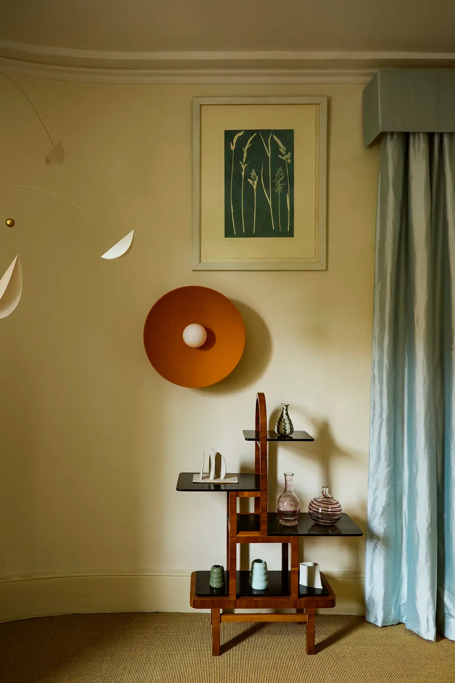 A styled interior corner with a light blue curtain, and a small wooden shelving unit holding decorative glass vessels. Above the shelf hangs a framed print and a round orange wall lamp.