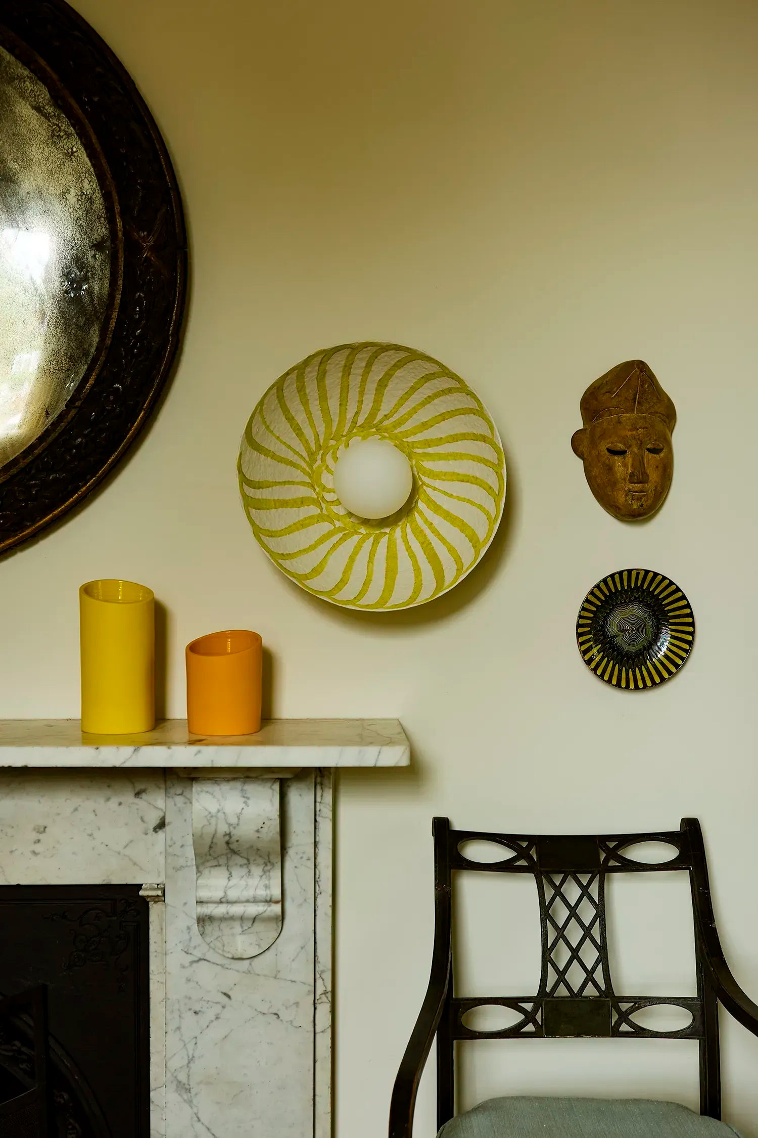 A styled interior scene with a cream wall, a marble mantel, a dark wooden chair, two yellow-orange candles, and several decorative wall pieces including a large yellow and white wall light with glass diffuser and a brown sculptural mask.