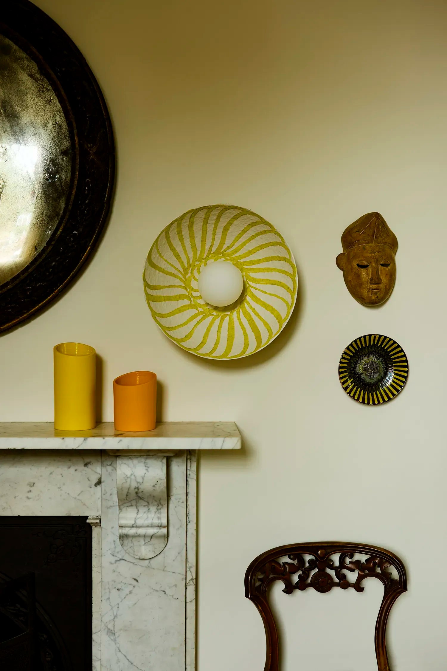 A styled interior scene showing decorative wall art and candles above a marble mantel. The central product is a yellow and white spiral-pattern wall light with glass diffuser.