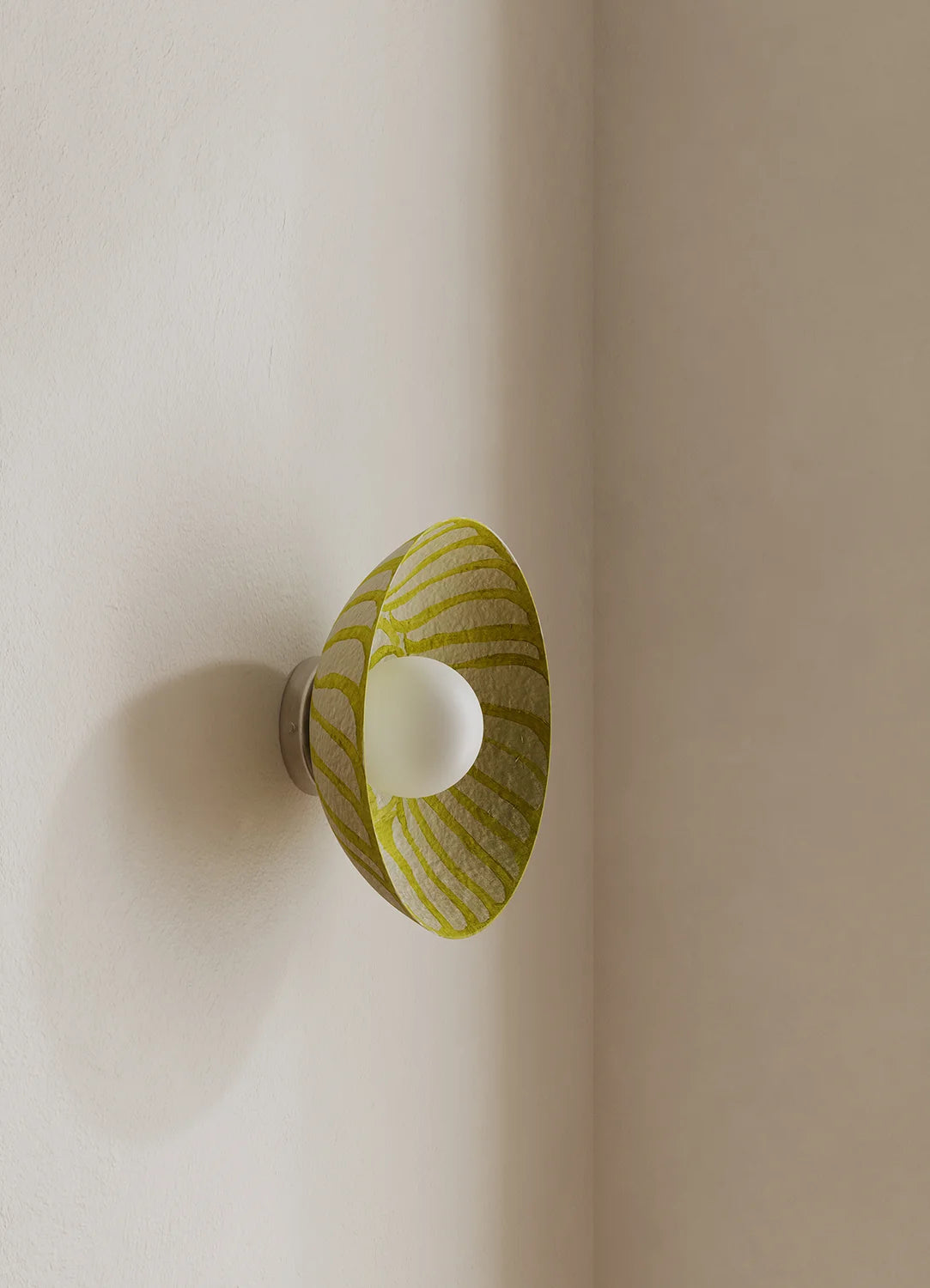 A modern wall-mounted sconce with a conical shade featuring a yellow and white striped pattern, casting a soft glow from a central white bulb, mounted on a beige wall.