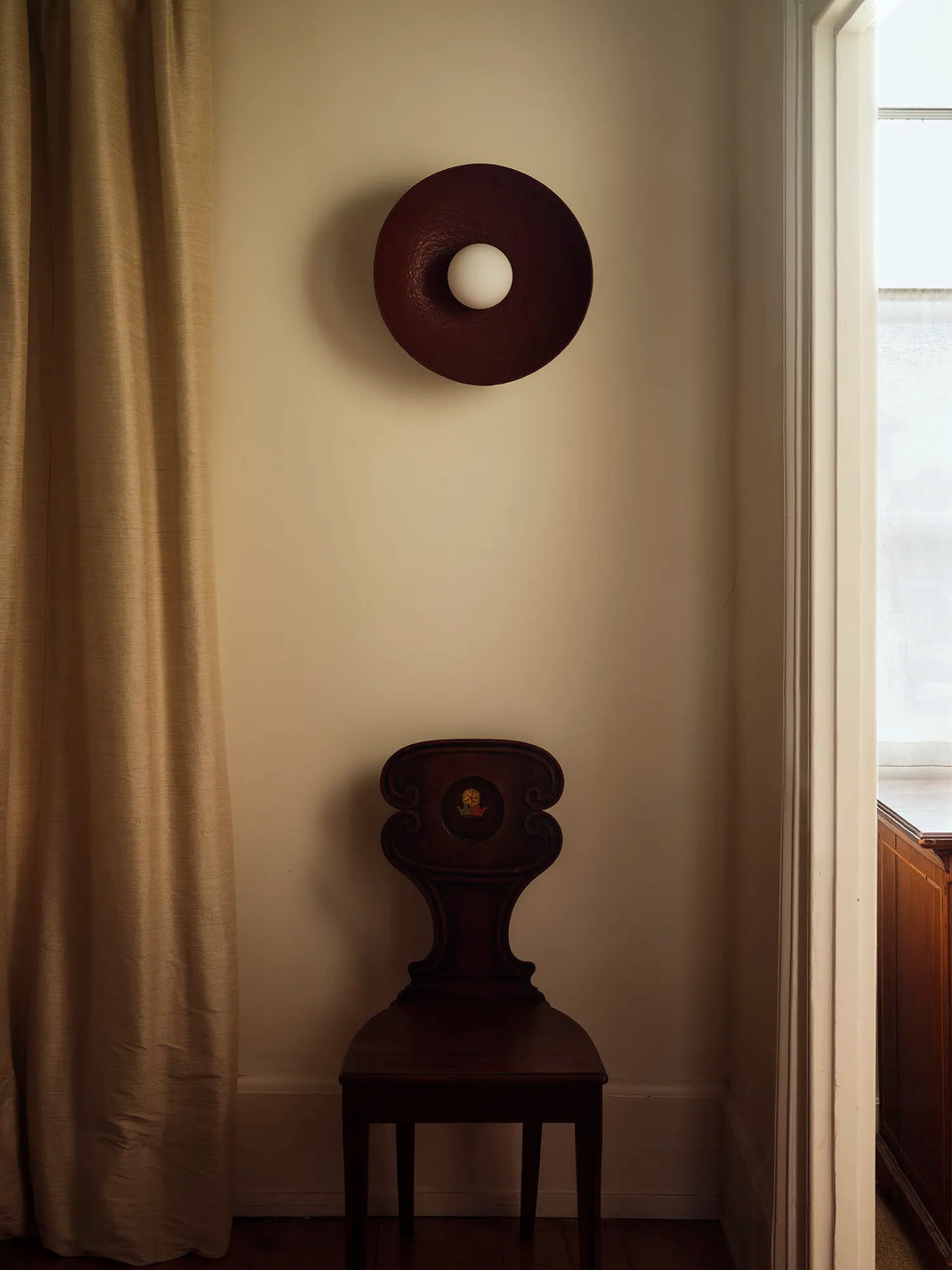 A dark red circular wall sconce with a white globe bulb, mounted on a beige wall above a dark wood chair in a room with a curtain.