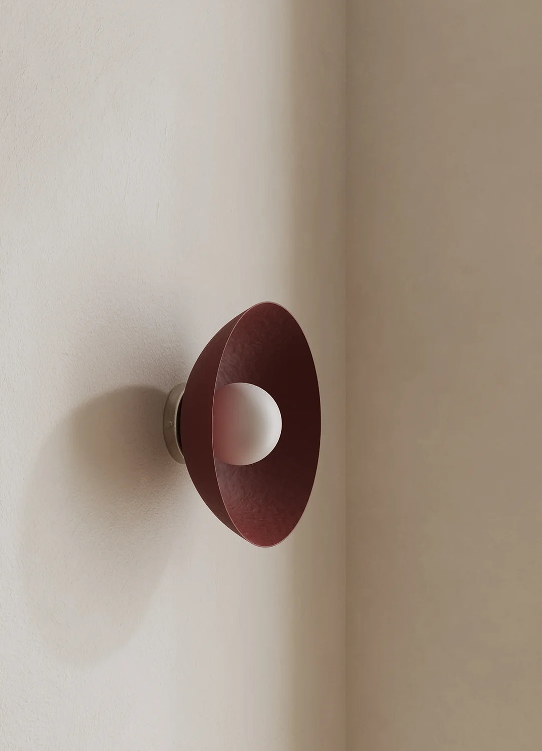 A modern wall-mounted light fixture with a dark red conical shade and a white spherical bulb, mounted on a beige wall in a corner.