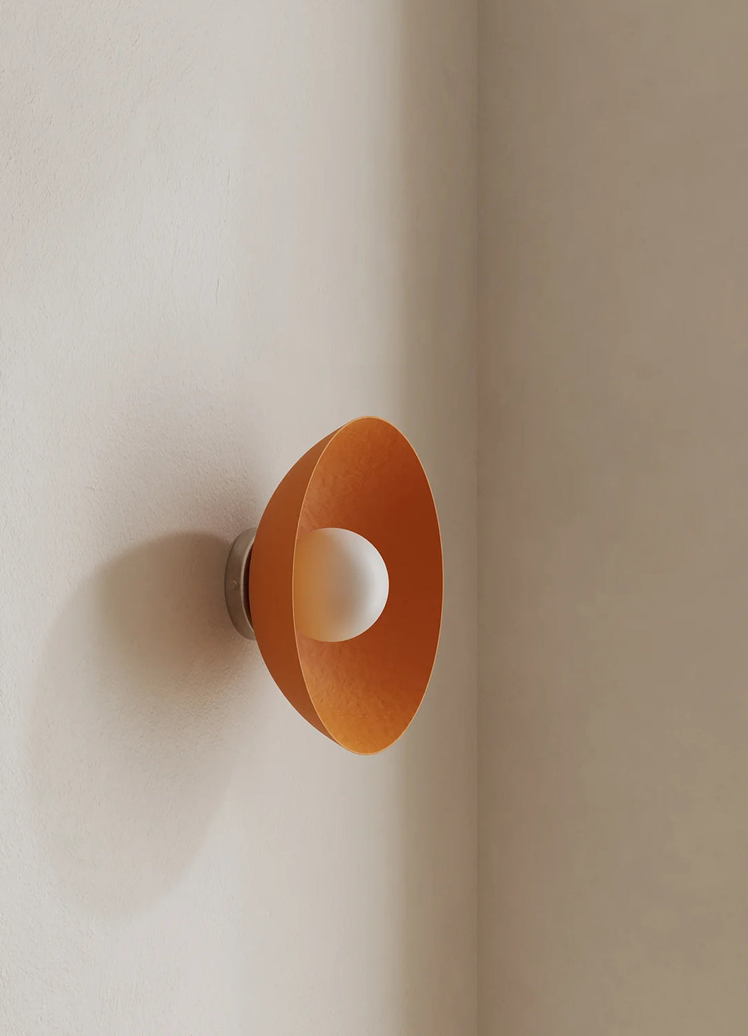 A modern wall-mounted light fixture with a wide, shallow orange dome shade and a white spherical bulb or diffuser at the center, mounted on a warm beige plaster wall.