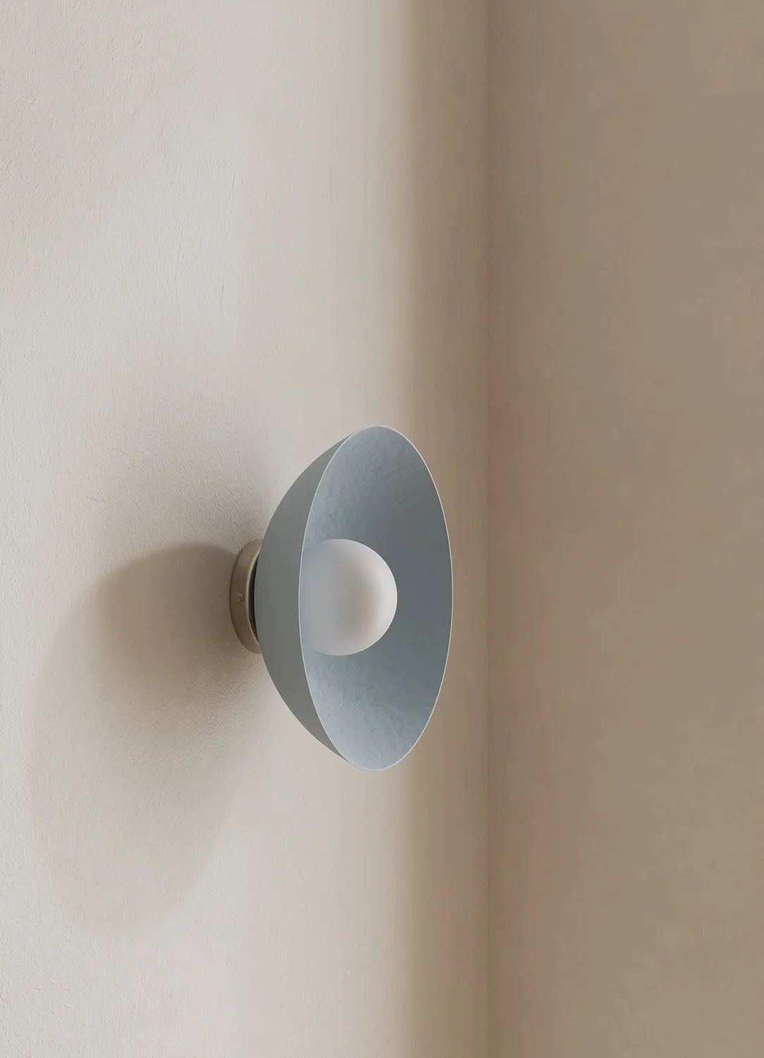 A modern wall-mounted light fixture with a wide, shallow shade in a pale blue finish and a frosted white bulb inside, mounted on a warm beige plaster wall.