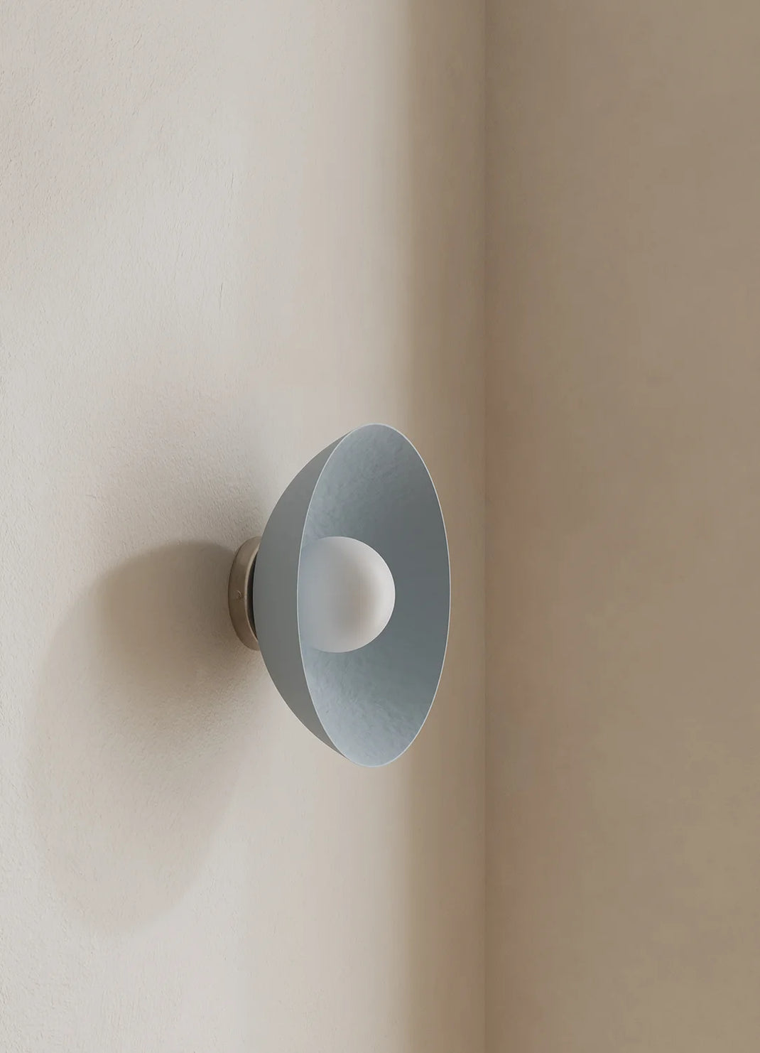 A modern wall-mounted light fixture with a wide, shallow shade in a pale blue finish and a frosted white bulb inside, mounted on a warm beige plaster wall.