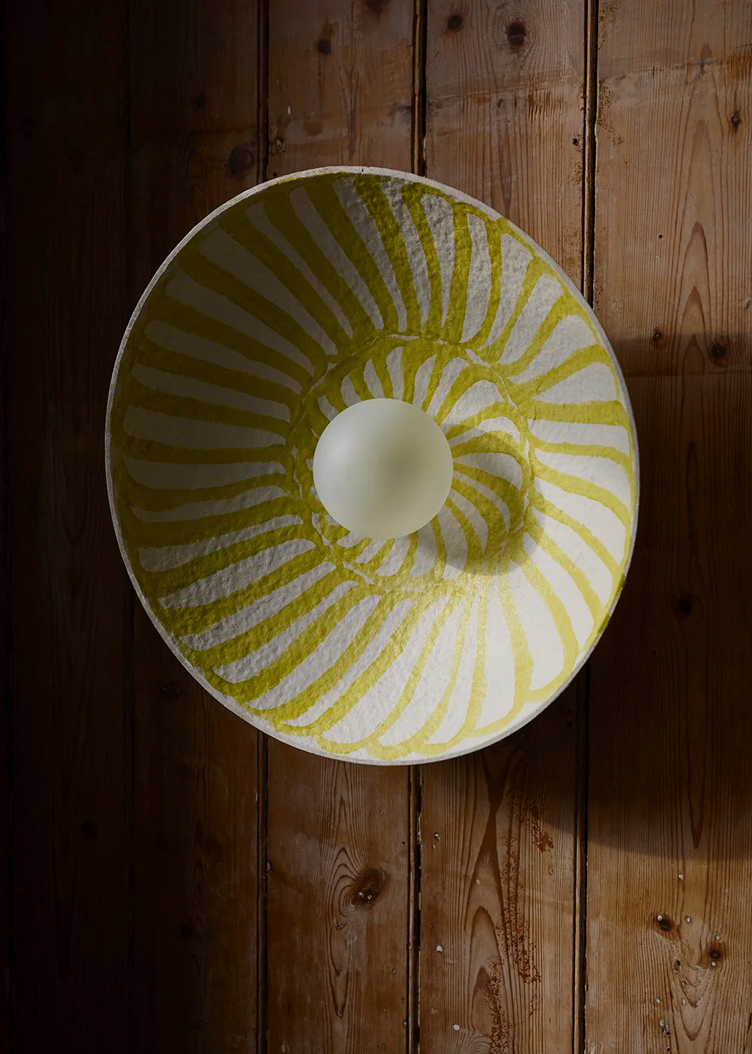 A large white wall lamp with a yellow-white sunburst pattern inside, photographed from above on a dark wood plank surface.