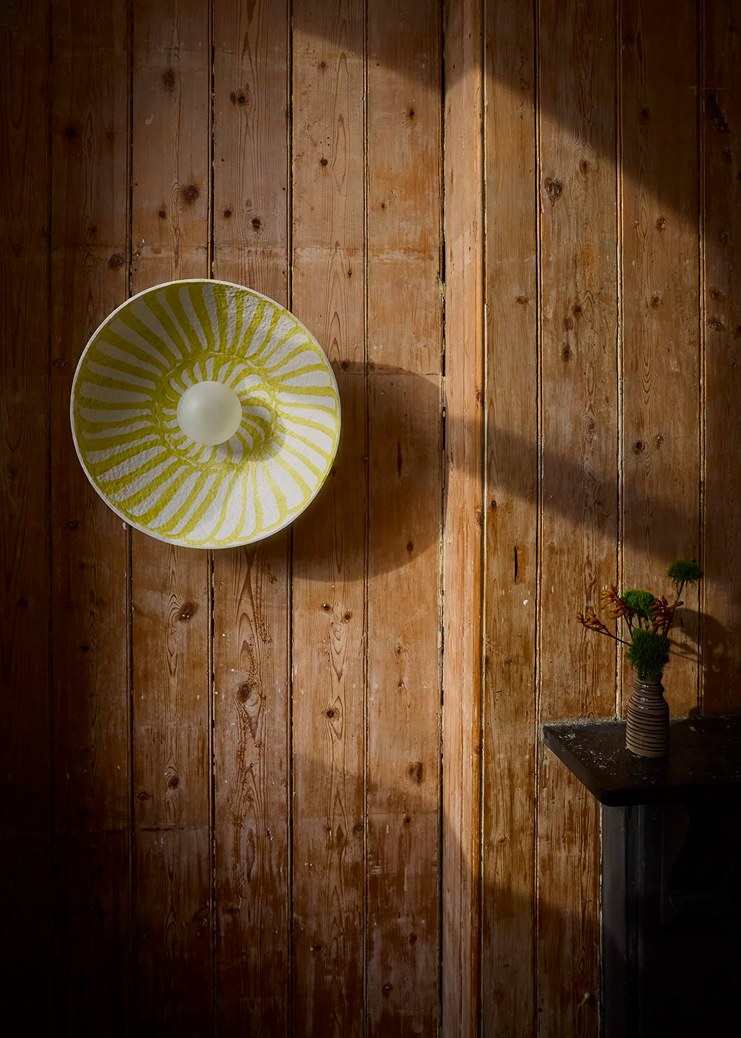 A round wall light with a yellow and white sunburst pattern, mounted on a rustic wooden plank wall next to a small table with a vase of flowers.