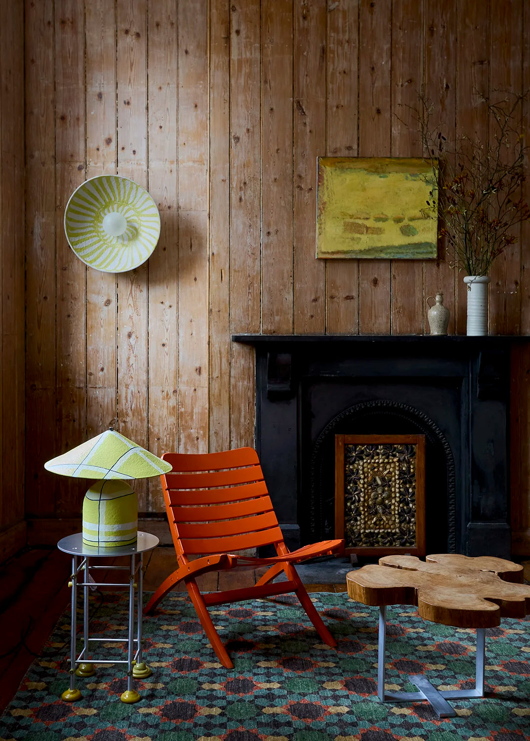 A styled interior scene with a wood-paneled wall, black fireplace, patterned rug, orange slatted chair, small side table with a yellow-and-white lamp, and a flower-shaped wooden table.