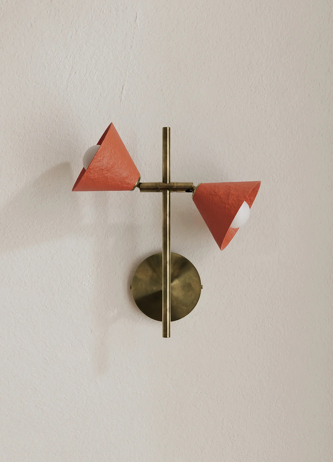 A modern wall-mounted light fixture with a brass-finished metal frame and two cone-shaped shades in a terracotta orange-red color, mounted on a plain off-white wall.