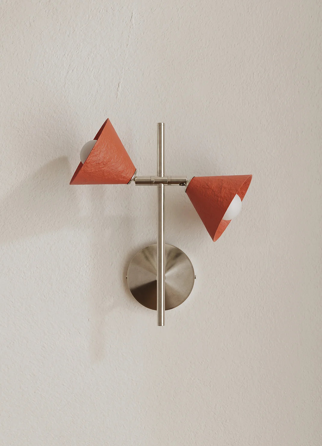 A modern wall-mounted light fixture with two terracotta-colored cone-shaped shades and a brushed silver metal base and central rod, mounted on a textured off-white wall.