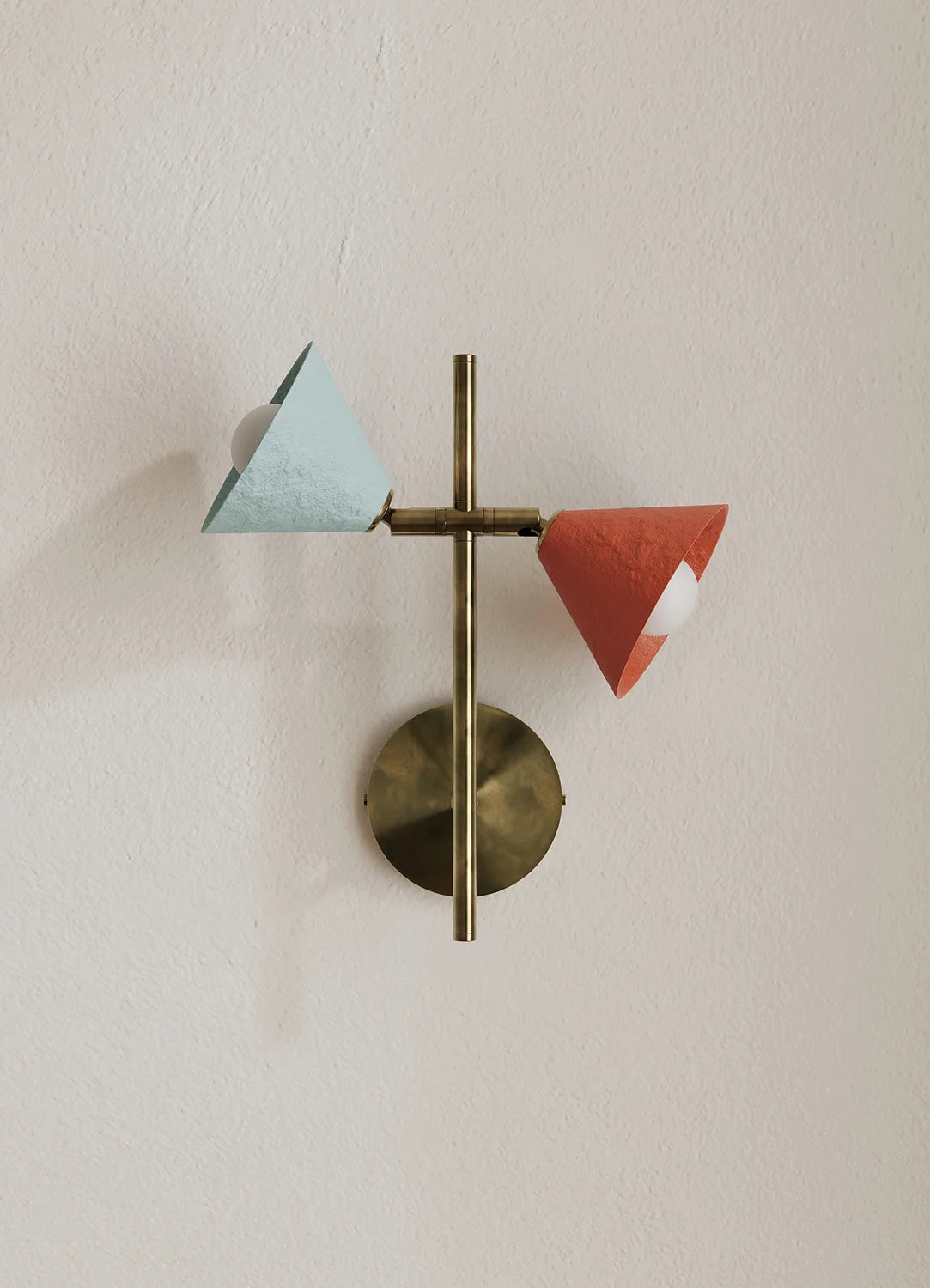 A modern wall-mounted light fixture with a brass circular backplate and vertical rod, featuring two angled paper-like shades in pale blue and terracotta red against a warm off-white wall.