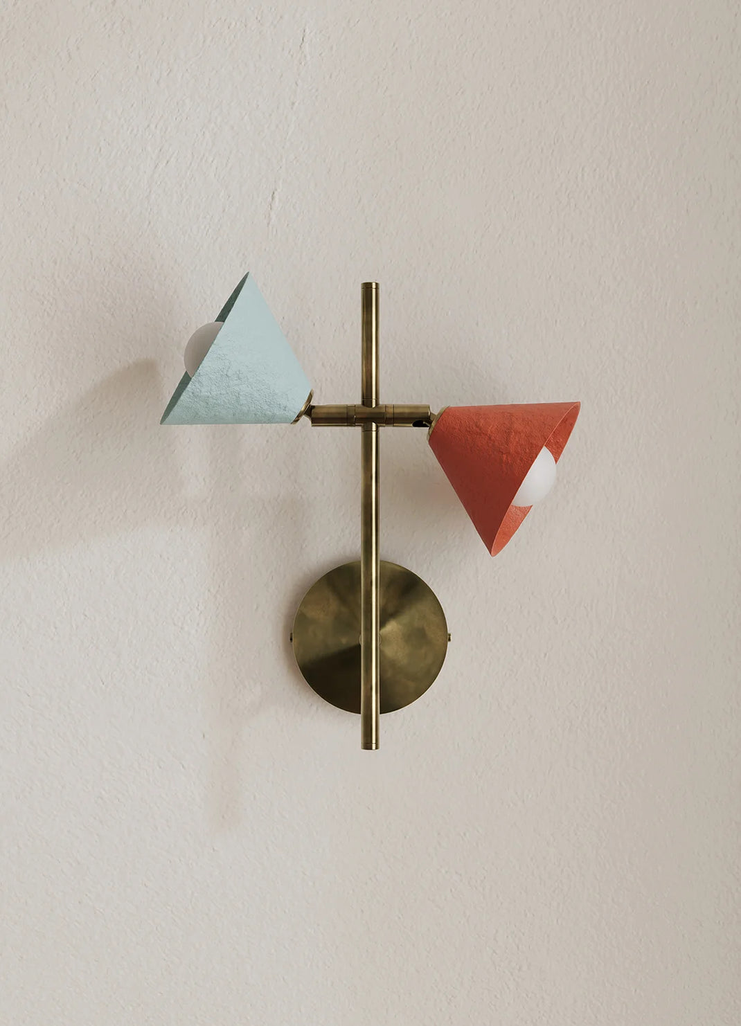 A modern wall-mounted light fixture with a brass circular backplate and vertical rod, featuring two angled paper-like shades in pale blue and terracotta red against a warm off-white wall.