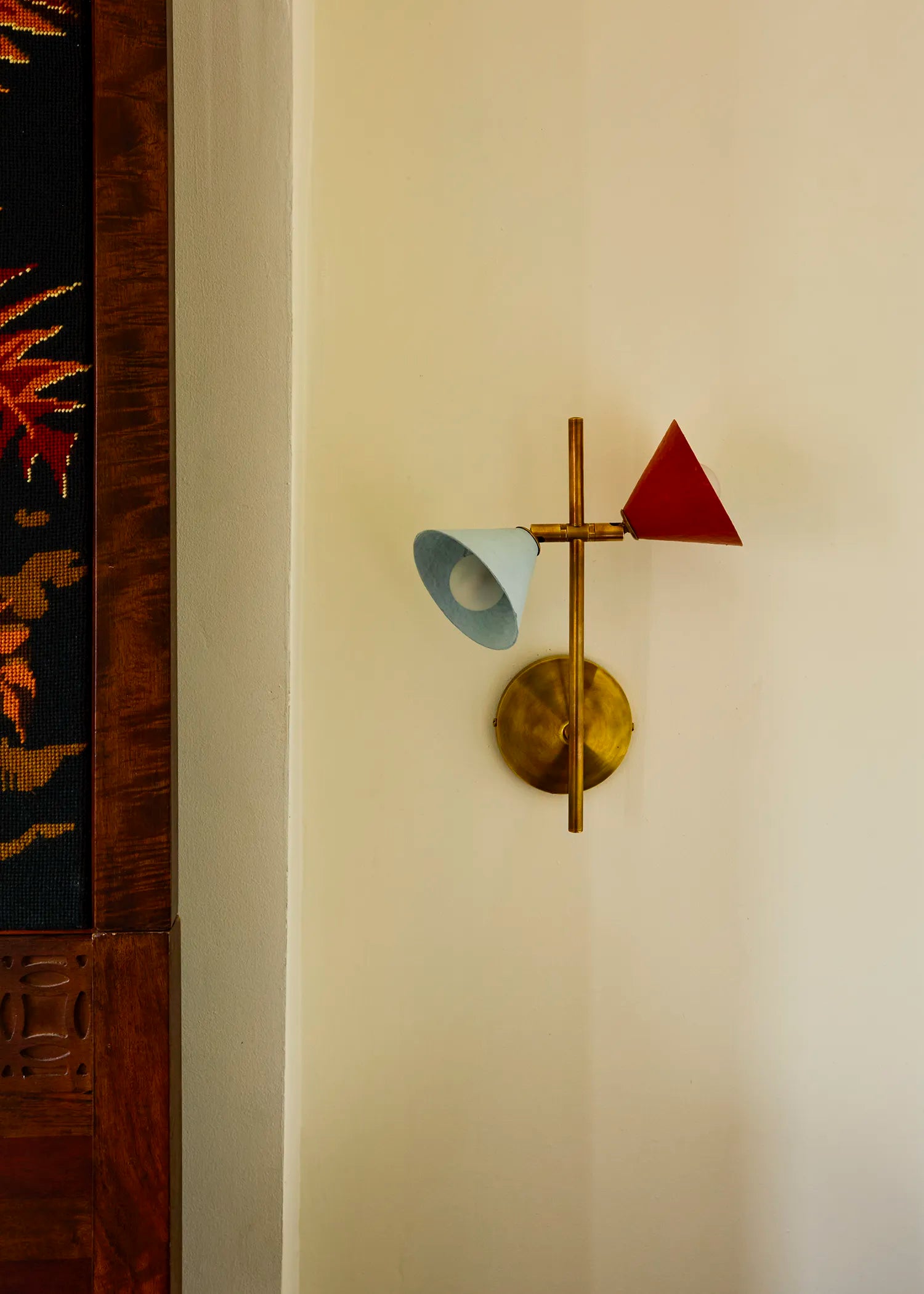 A modern brass wall sconce with a light blue and red cone shades, mounted on a cream-colored wall next to a dark wood panel.