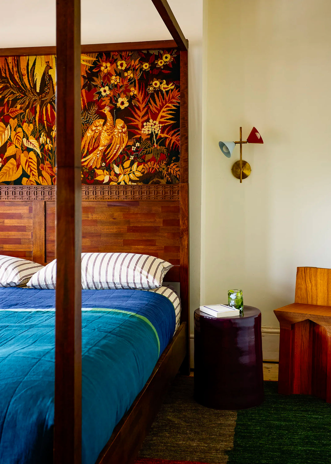 A bedroom interior featuring a wooden four-poster frame bed with a colorful tropical floral and bird tapestry above the headboard. A wall light with two different coloured shades sits above the dark side table.