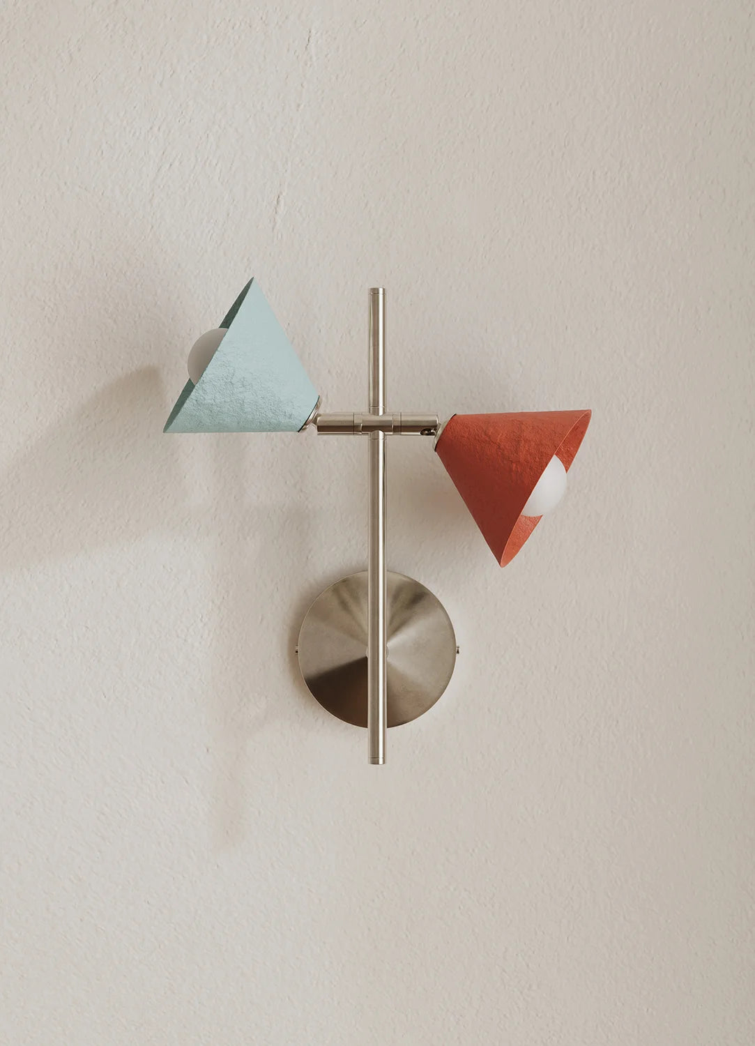 A modern wall-mounted light fixture with a brushed metal circular base and vertical rod, featuring two cone-shaped shades in pale blue and terracotta orange, set against a warm off-white textured wall.