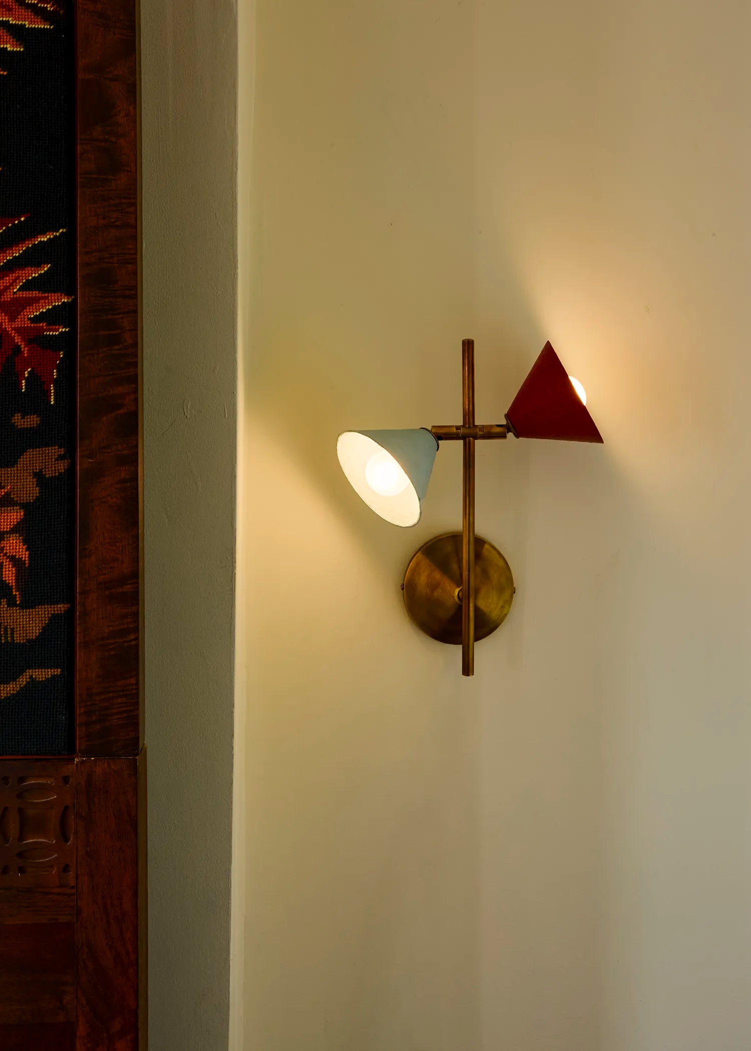 A modern wall sconce light fixture mounted on a cream-colored wall, featuring a brass-finished circular wall mount, a vertical brass rod, with one blue and one red conical shade.