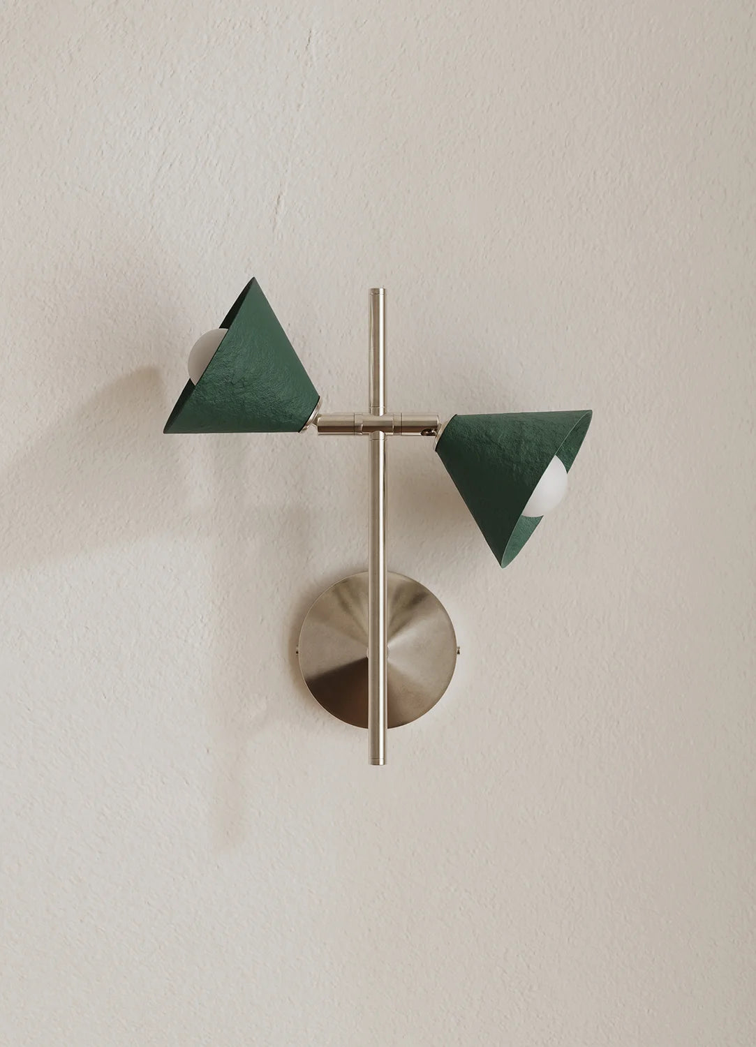 A modern wall-mounted light fixture with two dark green triangular shades and a brushed silver metal base and central rod, mounted on a textured off-white wall.