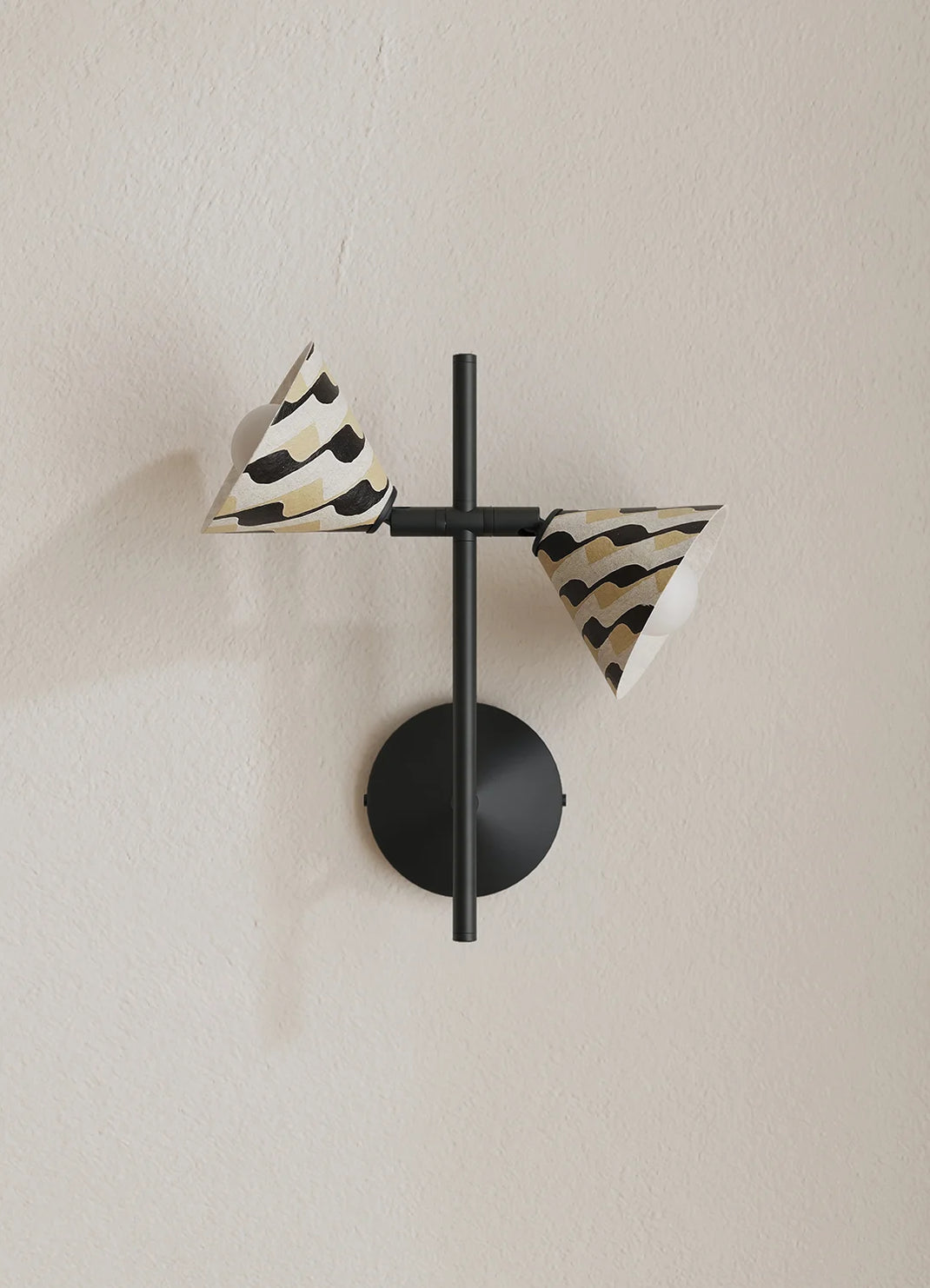 A modern wall-mounted light fixture with a matte black circular base and crossbar, featuring two cone-shaped shades with a black and beige zebra-like pattern, mounted on a textured beige wall.