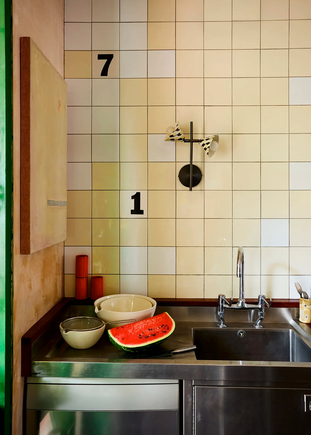 A kitchen scene with cream and beige square wall tiles, a stainless steel sink and countertop, a cut watermelon on the counter, and a wall light with a black and white pattern is attached to the tiles.
