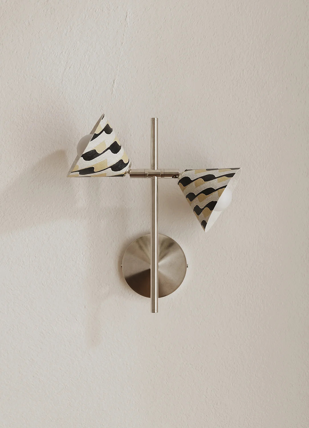 A modern wall-mounted light fixture with two cone-shaped shades featuring a black and beige wavy pattern, suspended from a brushed silver metal crossbar on a circular base against a textured off-white wall.
