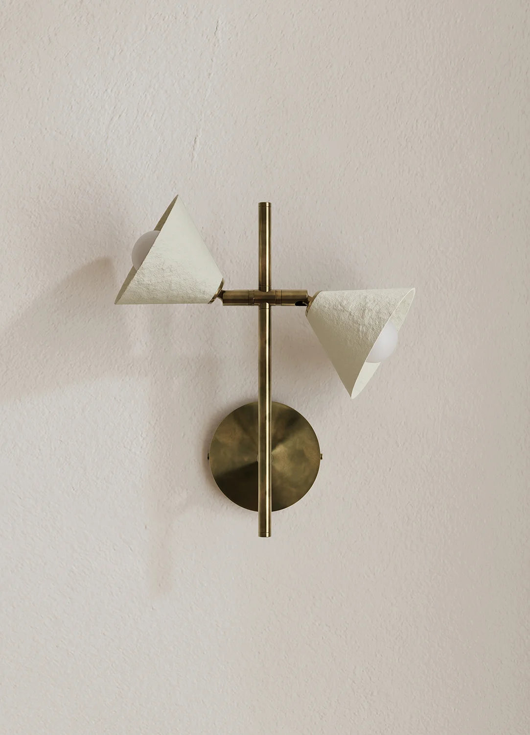 A modern wall-mounted light fixture with a vertical brass rod, a round brass base, and two angled white textured cone shades, shown against a plain off-white wall.