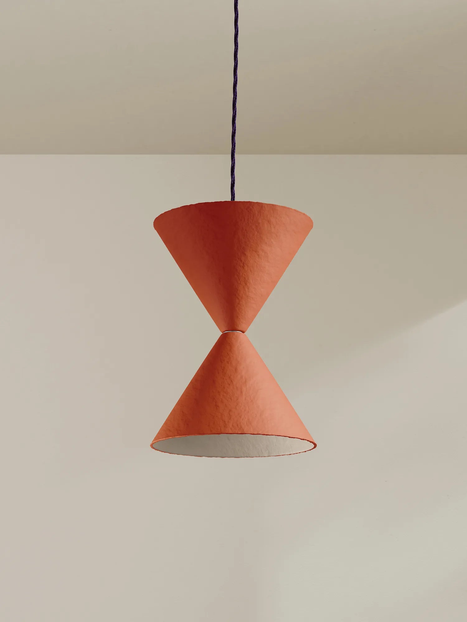 A modern pendant light with two textured terracotta-colored cone shades connected by a thin dark blue cord, hanging against a minimalist beige wall and ceiling.