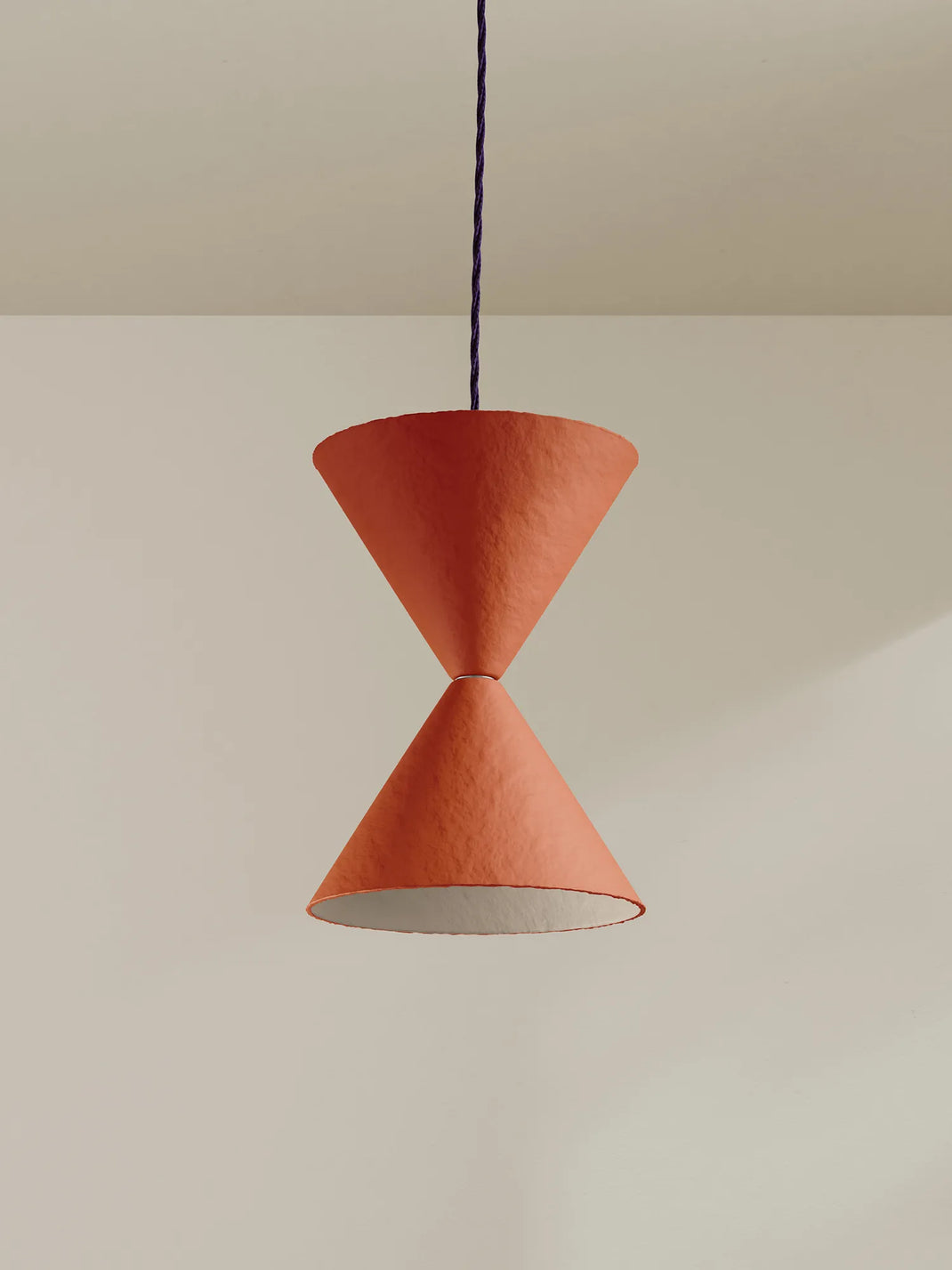 A modern pendant light with two textured terracotta-colored cone shades connected by a thin dark blue cord, hanging against a minimalist beige wall and ceiling.