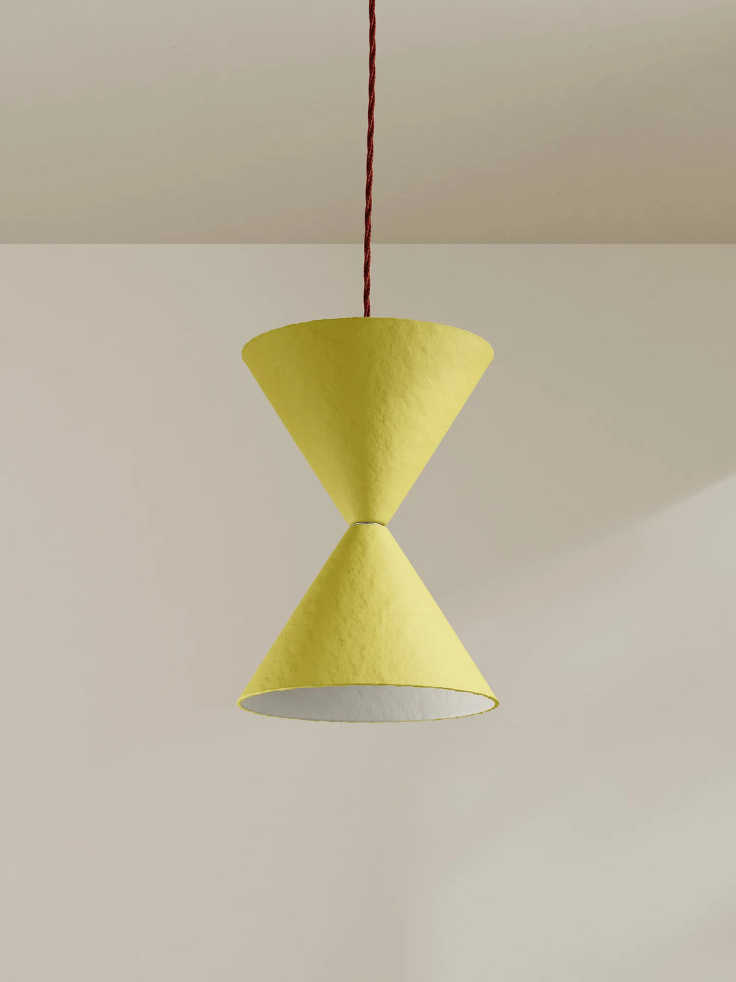 A modern pendant light with two textured yellow cone shades connected at the center, suspended by a burgundy cord against a minimalist beige wall and ceiling.