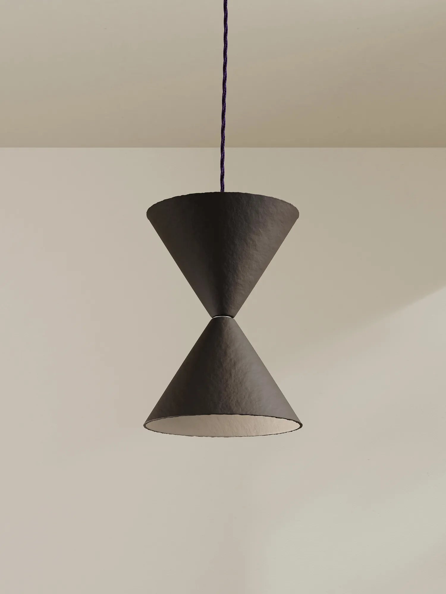 A modern pendant light with two textured brown-black cone shades connected at the center, suspended by a dark blue cord against a warm beige wall.