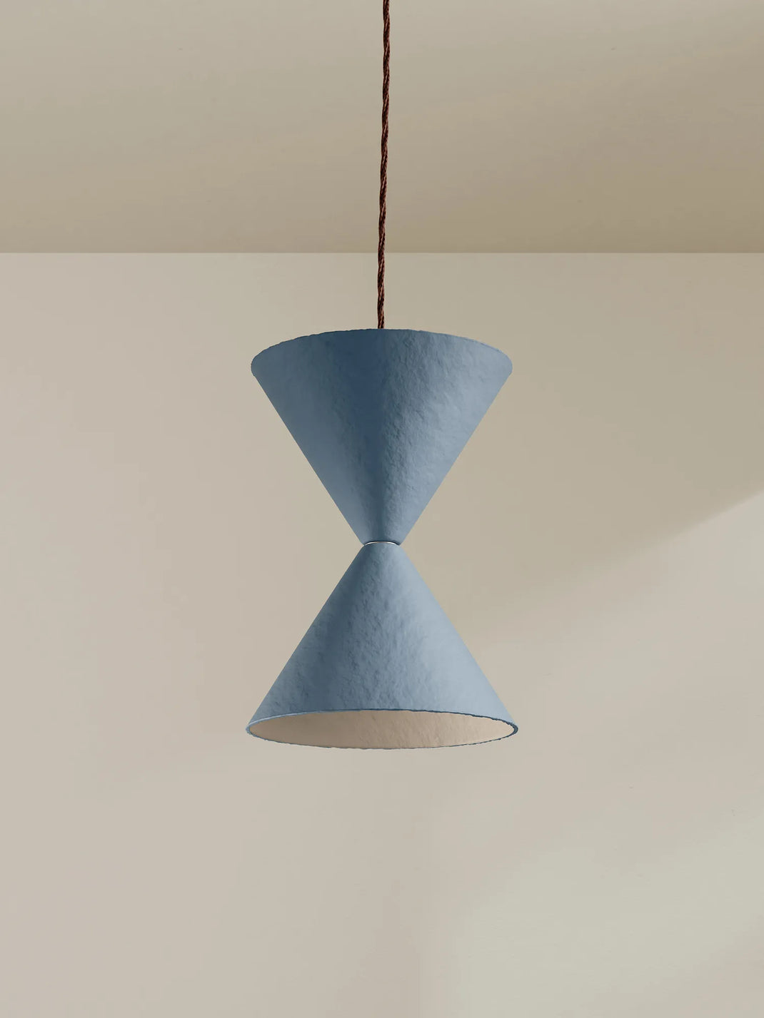 A modern pendant light with two textured blue paper cone shades connected by a dark brown hanging cord, shown against a minimalist beige wall.