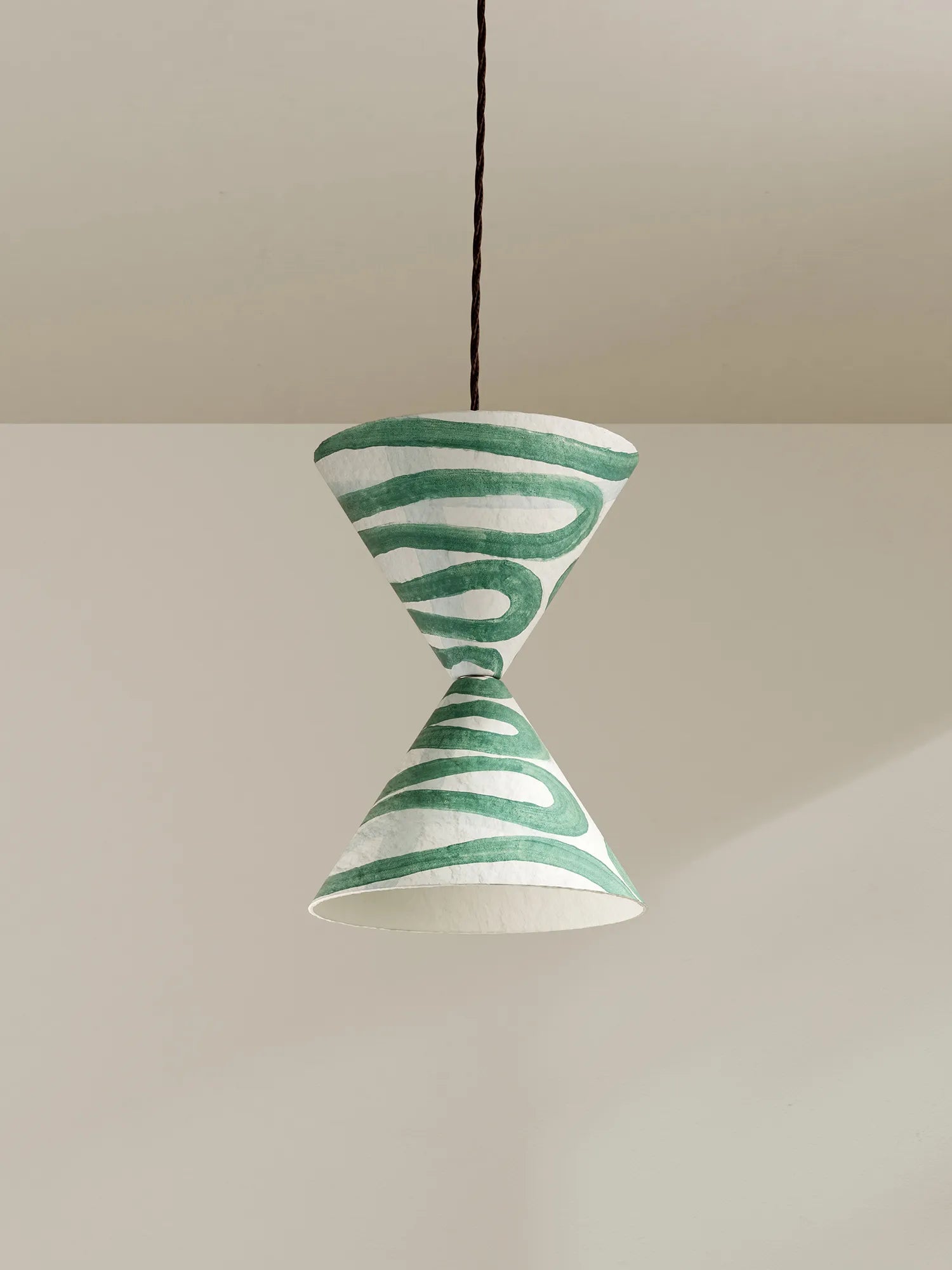 A sculptural pendant light with a white paper cone shade featuring green hand-painted wavy stripes, suspended by a brown cord against a minimalist beige background.