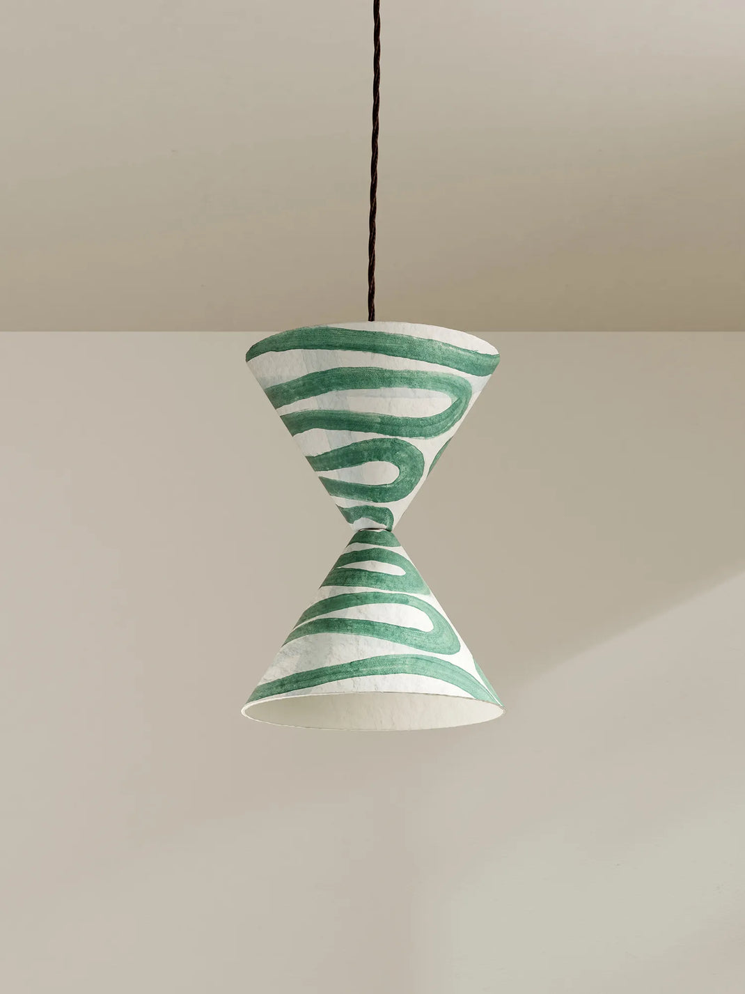 A sculptural pendant light with a white paper cone shade featuring green hand-painted wavy stripes, suspended by a brown cord against a minimalist beige background.
