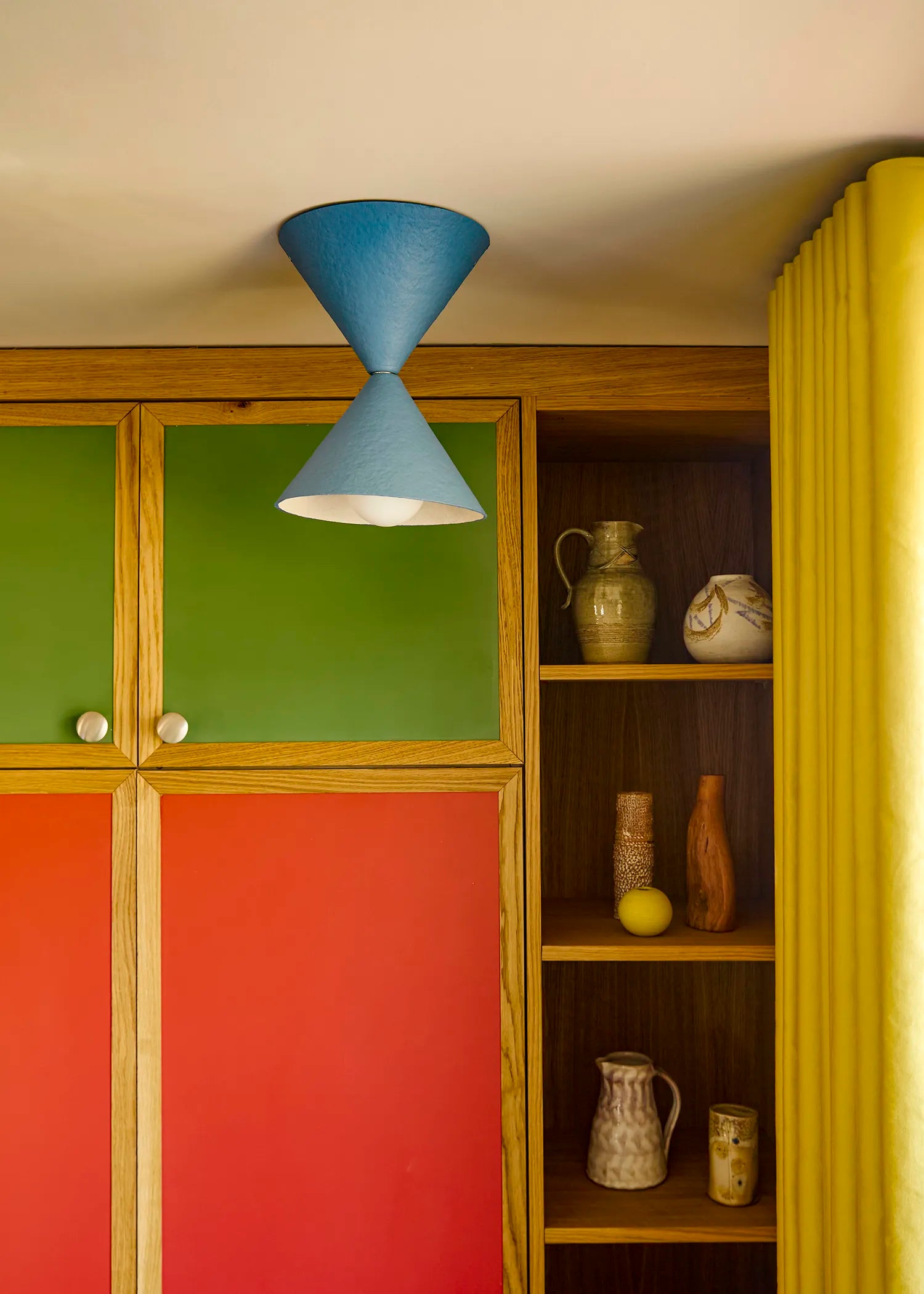 A mid-century modern dual-cone pendant light in blue hangs above a colorful wooden cabinet with green, red, and yellow doors, next to an open shelf displaying ceramic vases.