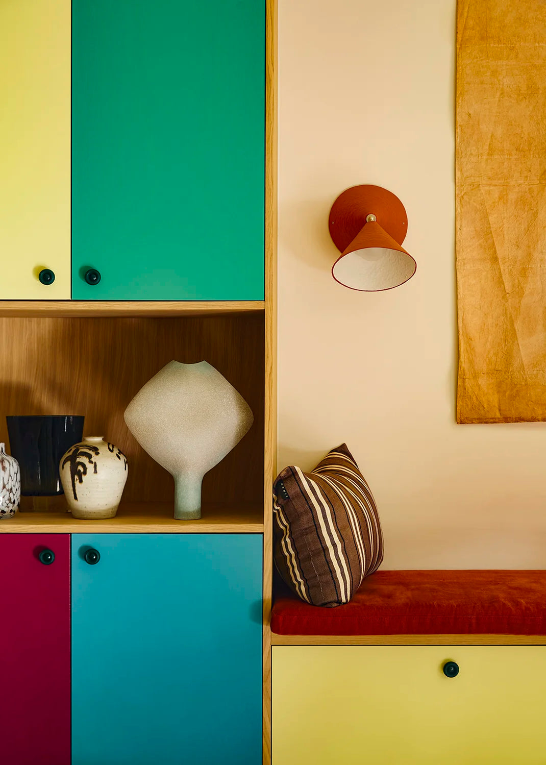 A colorful wooden storage cabinet with bright teal, yellow, magenta, and brown doors, styled with ceramic vases and a striped cushion in a warm interior setting. A red-brick wall sconce is mounted above the seating area.