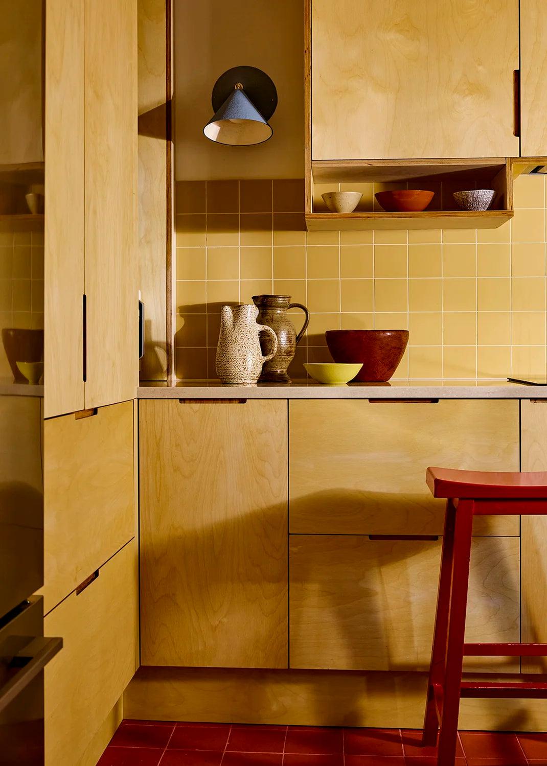 Kitchen with wooden cabinets, yellow tiles, and a red stool. A blue sconce is mounted on the wall.