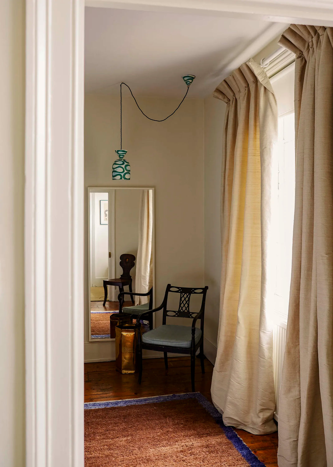 A view through a doorway into a room with beige curtains, a hanging pendant light, a black chair with a light blue seat, and a brown rug on a wooden floor.