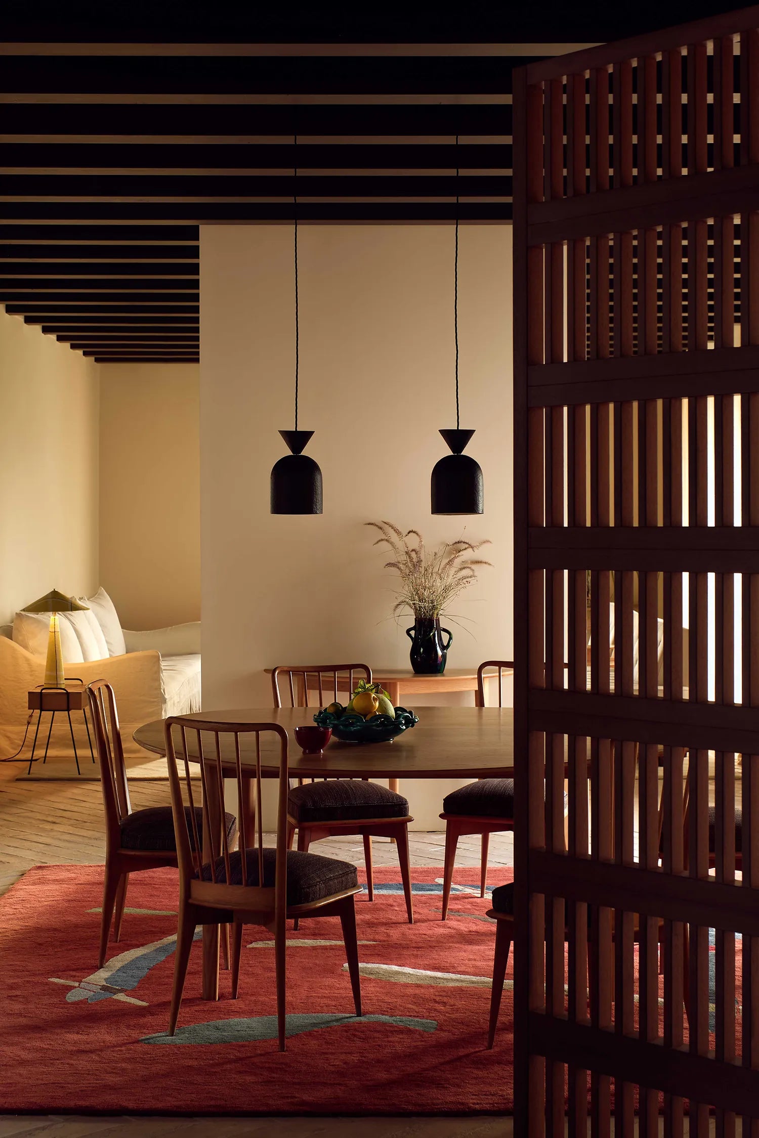 Dining room with wooden table and chairs, red rug, and decorative elements. Two dark-brown pendants hang above the dining table.