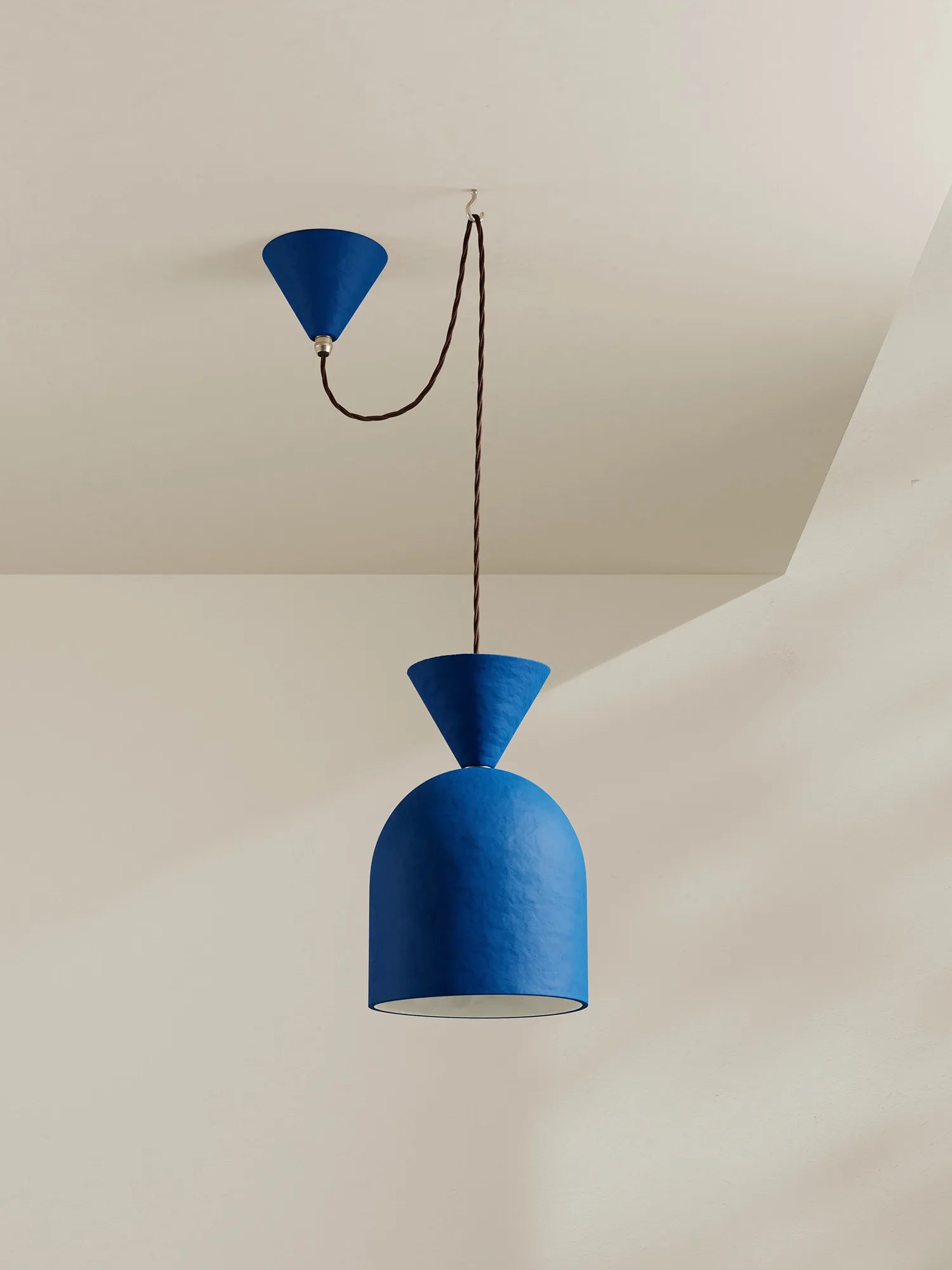 A modern blue pendant light with a textured blue dome shade, a conical blue cap, and a dark brown hanging cord, shown against a warm off-white ceiling and wall.