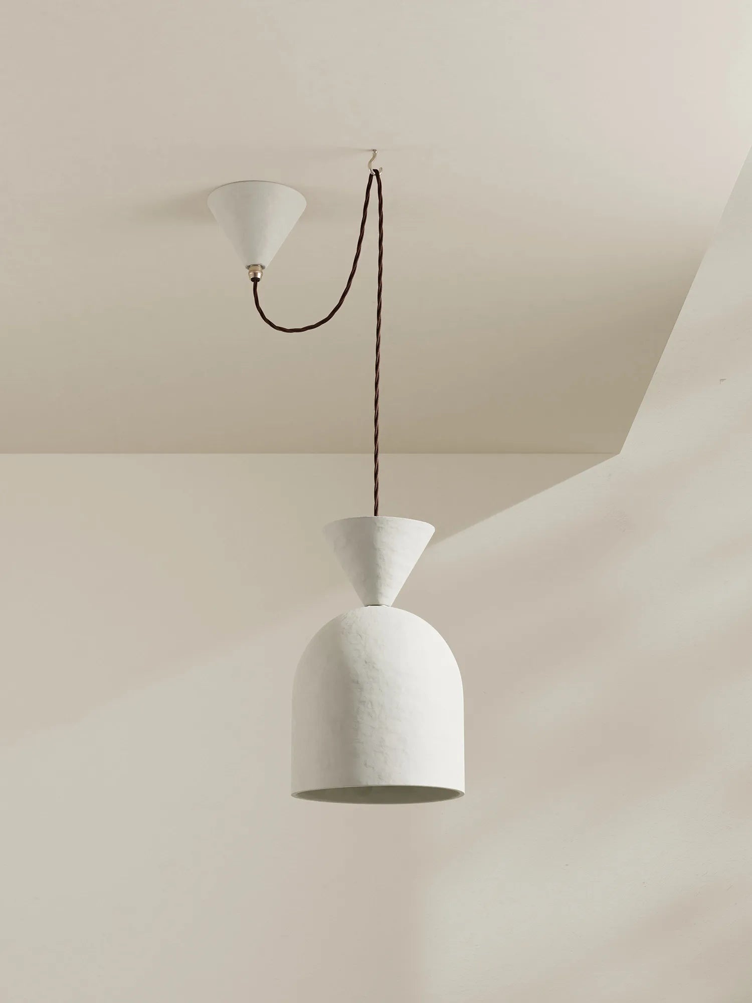 A minimalist white pendant light with a textured dome shade and a brown cord, suspended from a matching white ceiling canopy against a warm beige wall.