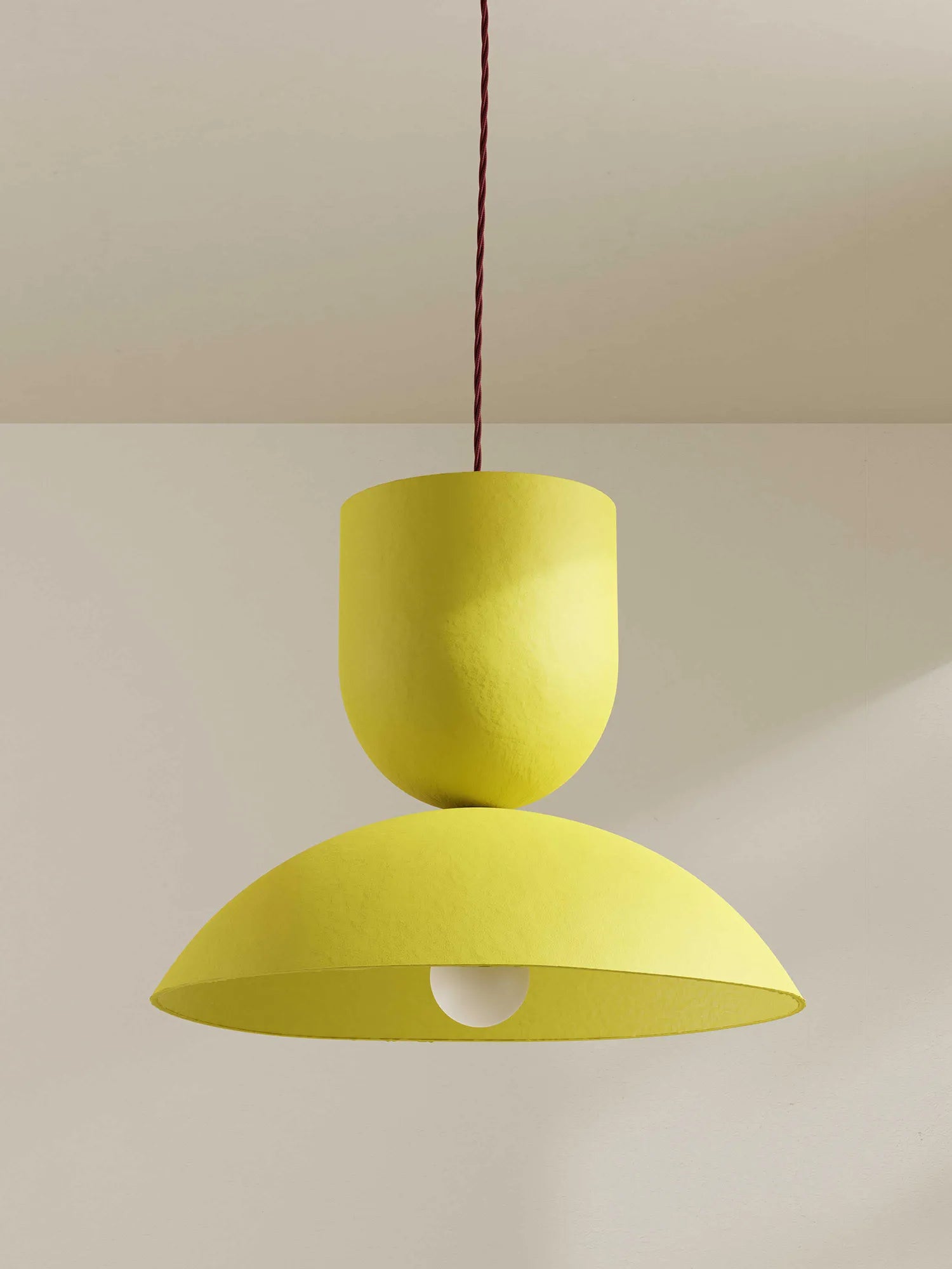 A modern two-tiered pendant light in a textured bright yellow finish, suspended by a dark red cord against a neutral beige wall.