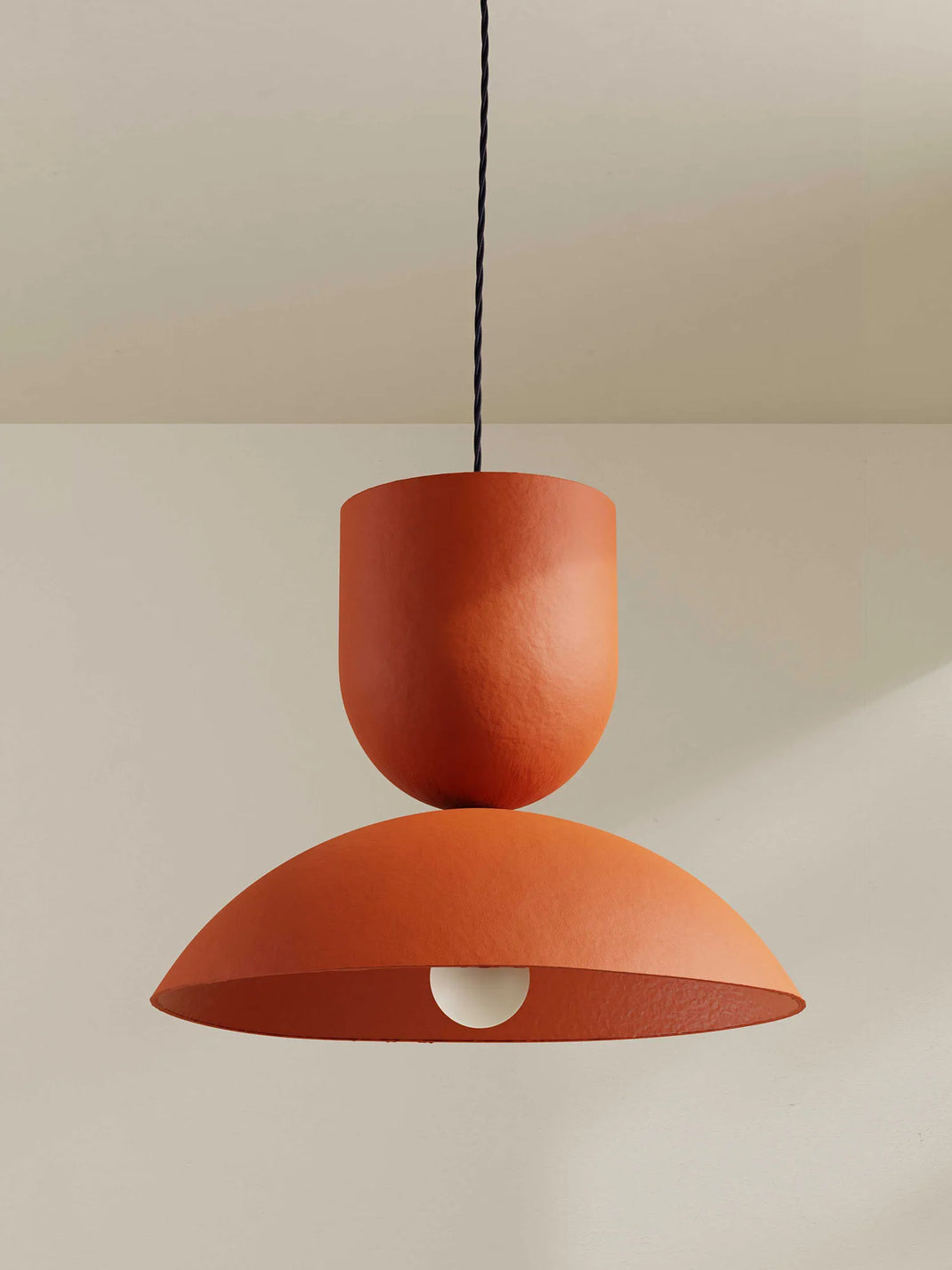 An orange terracotta pendant light with a two-tiered design, suspended by a midnight blue cord against a neutral beige wall.