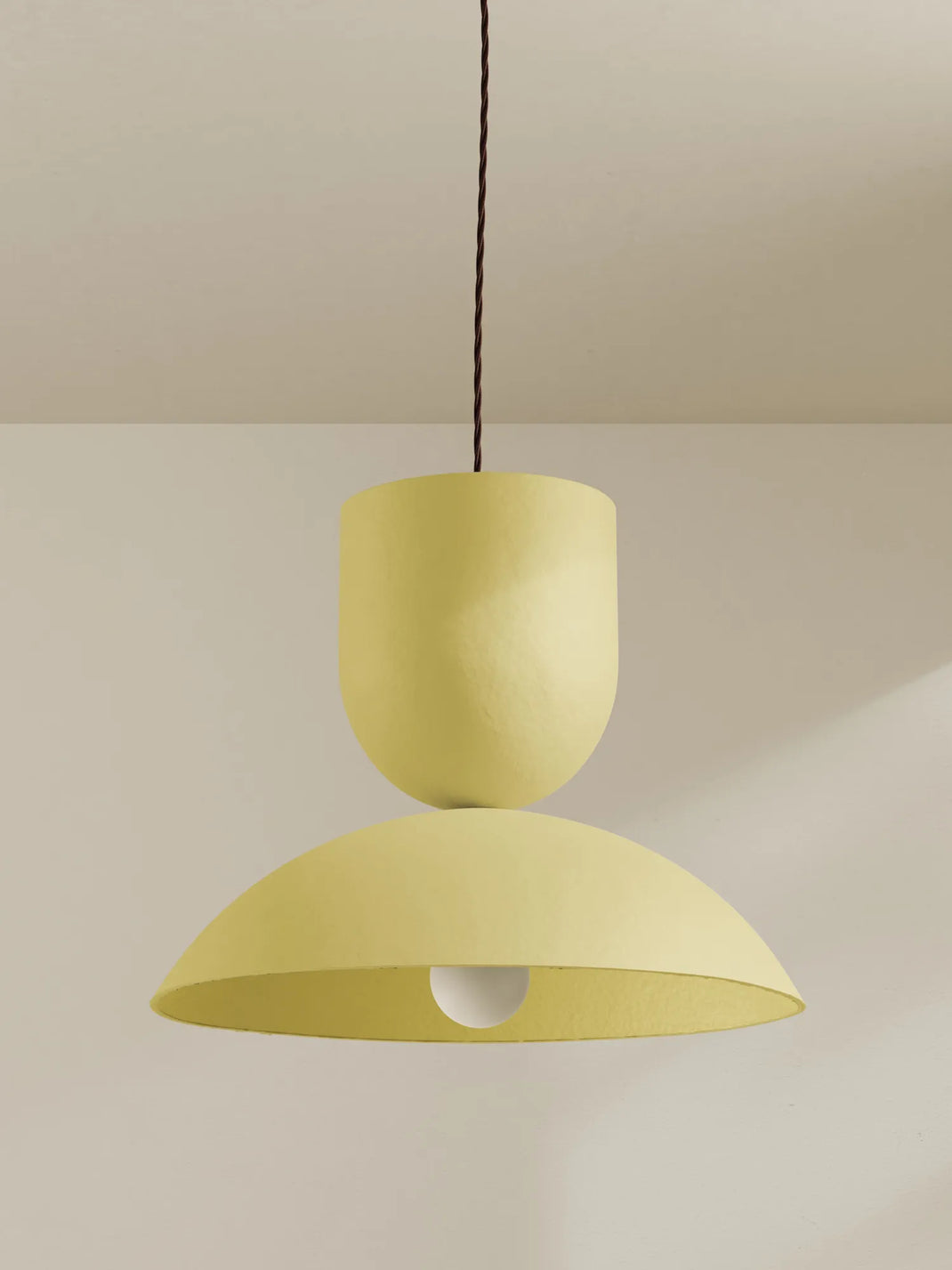 A modern two-tiered pendant light in a pale yellow color, suspended by a brown cord against a neutral beige wall and ceiling.