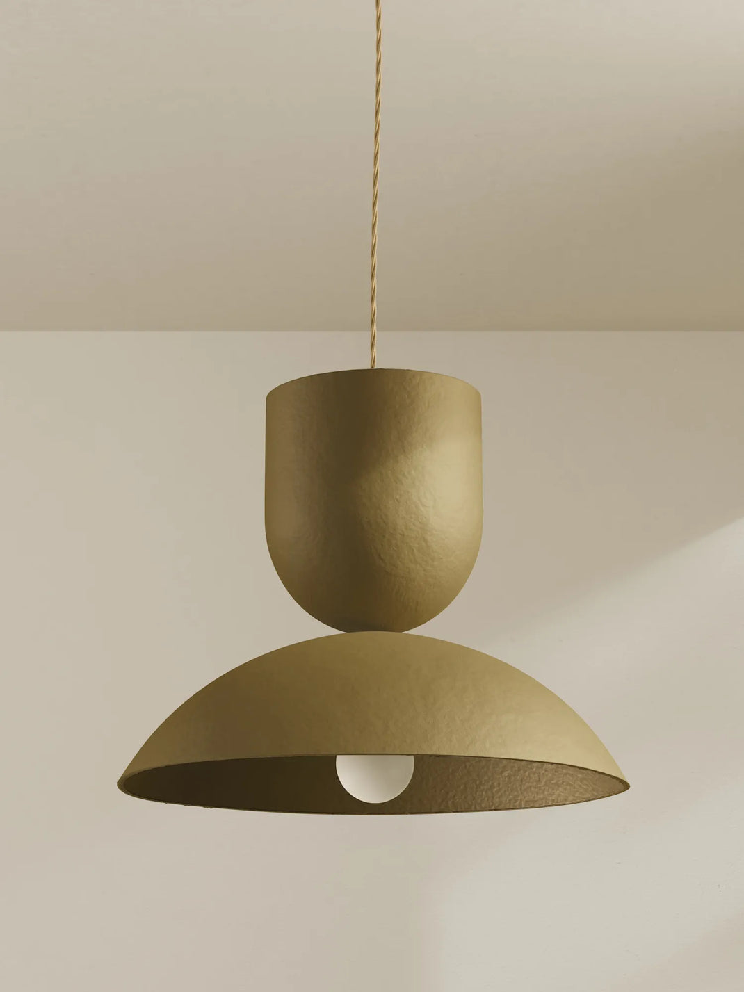 A modern pendant light with a textured finish in a warm moss color, featuring a stacked design with a cylindrical upper shade and a wide dome lower shade, suspended by a twisted cord against a neutral beige wall and ceiling.