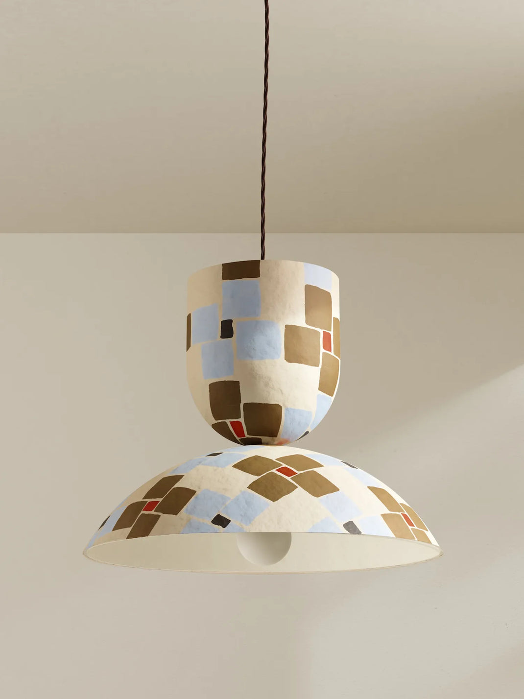 A modern two-tier pendant light with a sculptural, textured ceramic-like shade featuring a geometric checkered pattern in brown, beige, light blue, and red, suspended by a brown cord against a neutral beige wall.