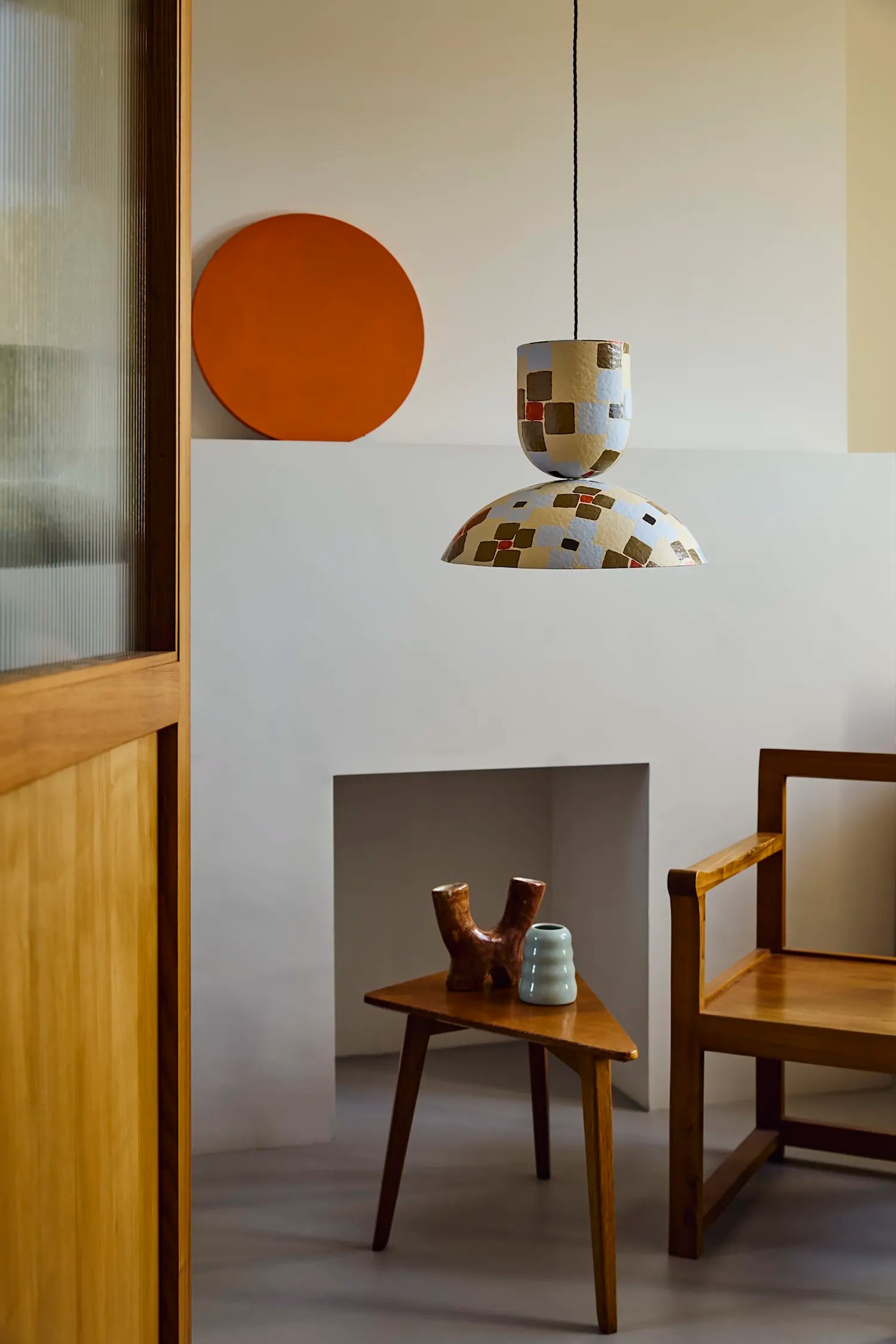 A modern pendant light with a white ceramic shade decorated with brown and gold geometric tiles, hanging in a minimalist room with a wooden chair, side table, and orange wall decor.